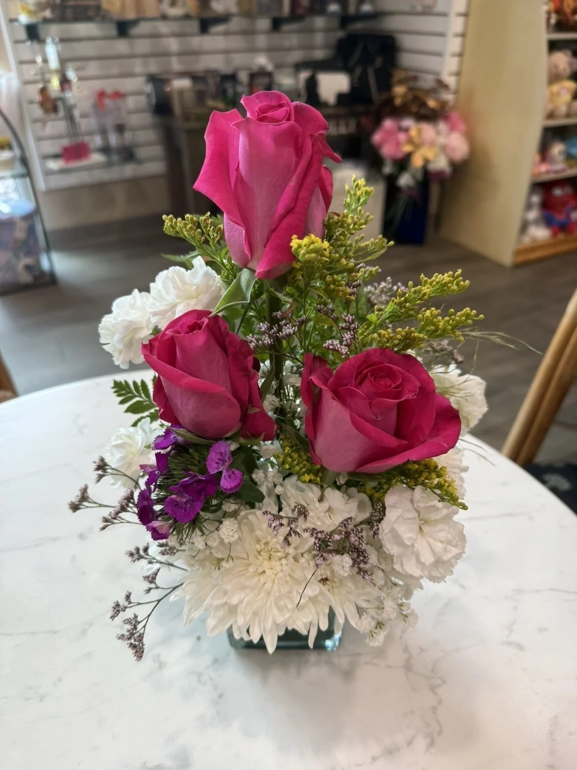 Un arrangement floral avec des roses roses roses, des fleurs blanches, à la lavande et des petites fleurs violettes dans un vase en verre sur une table blanche.