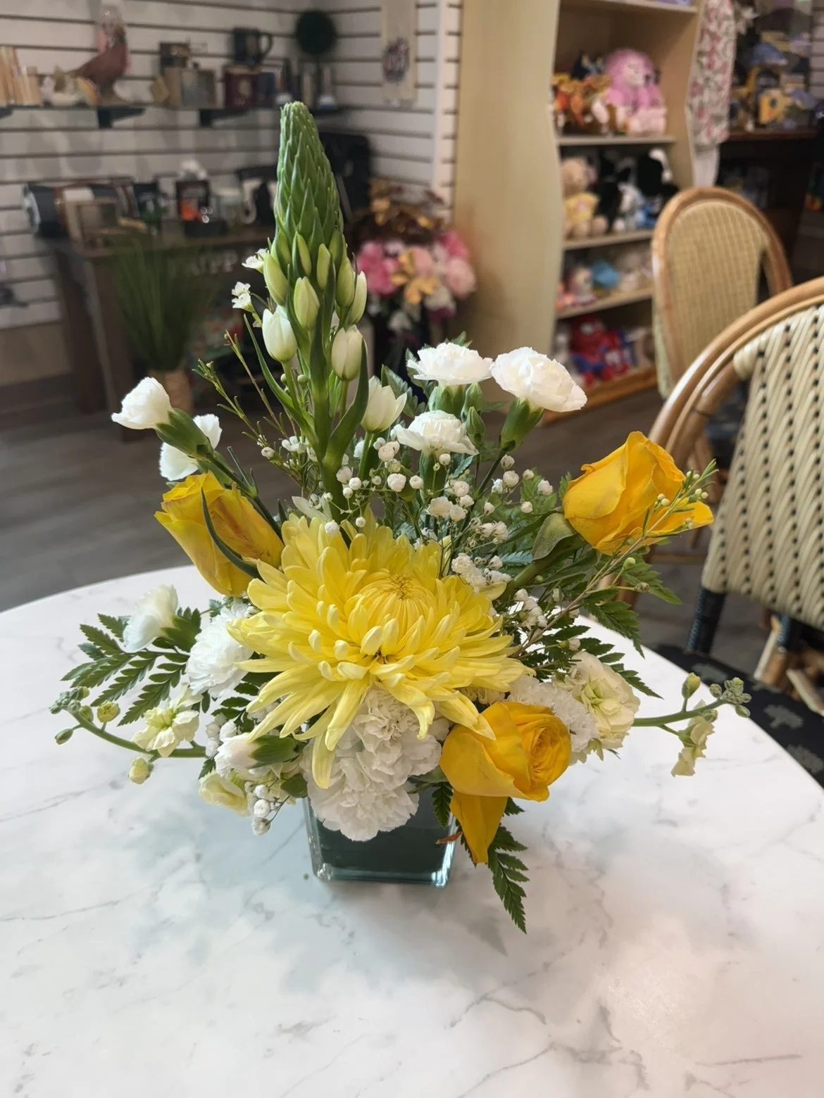 Un arrangement floral avec des fleurs jaunes, blanches et orange dans un vase en verre, posé sur une table blanche. En arrière-plan, une salle avec des étagères contenant des objets divers et des fauteuils en rotin.
