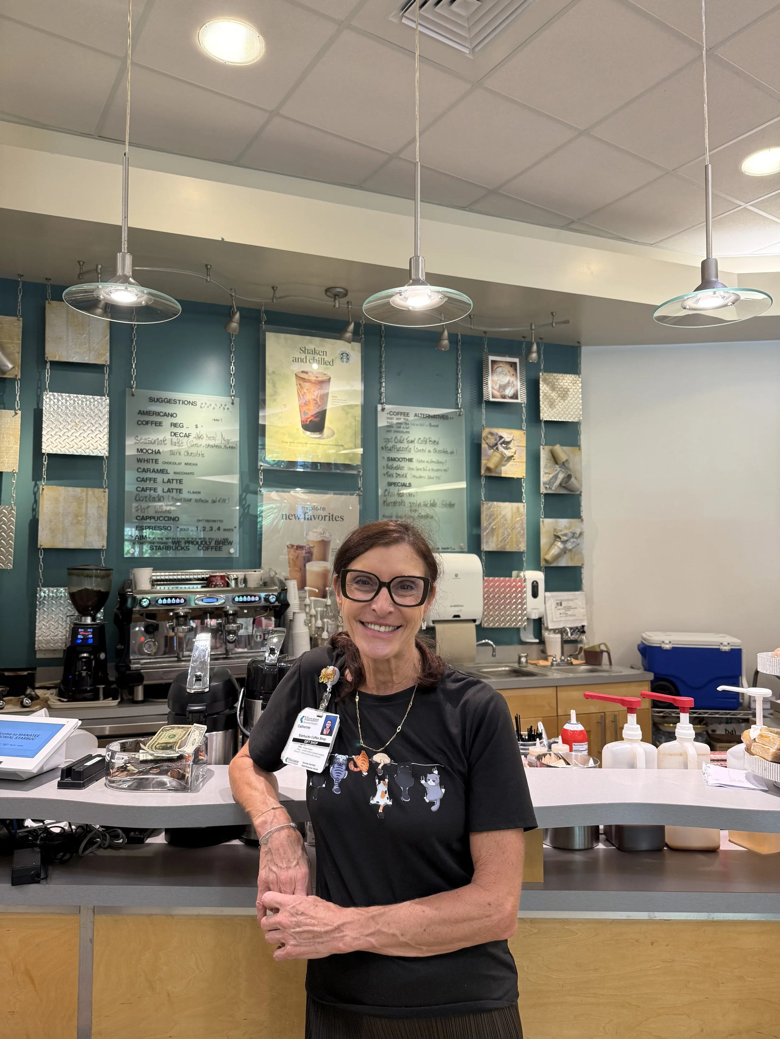 Une femme souriante portant des lunettes, travaillant au comptoir d'un café ou d'une boutique. Elle porte un badge et une chemise noire avec un motif de chiens. Derrière elle, un mur peint en bleu avec des menus et des décorations métalliques, une ma