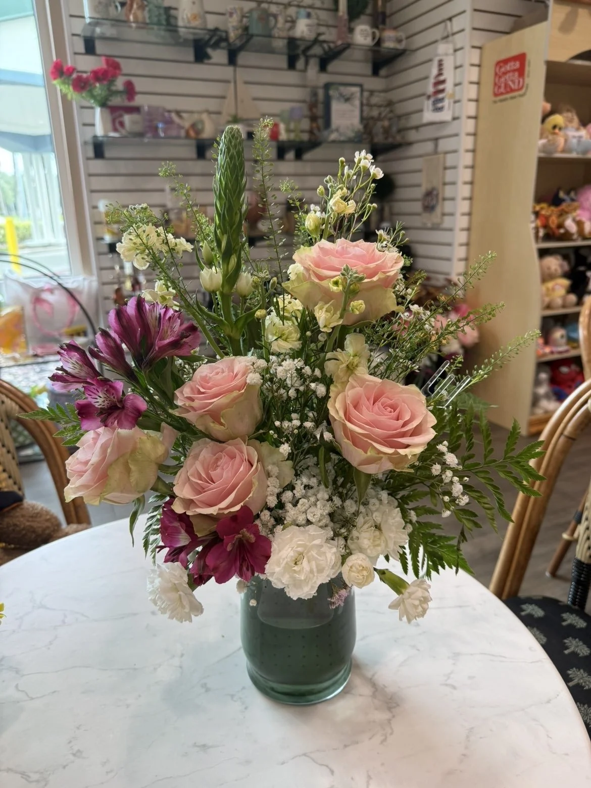 Un bouquet de fleurs dans un vase vert clair sur une table en marbre, avec des roses roses, des alstroemerias violettes et des petits fleurs blanches, dans un intérieur décoratif.