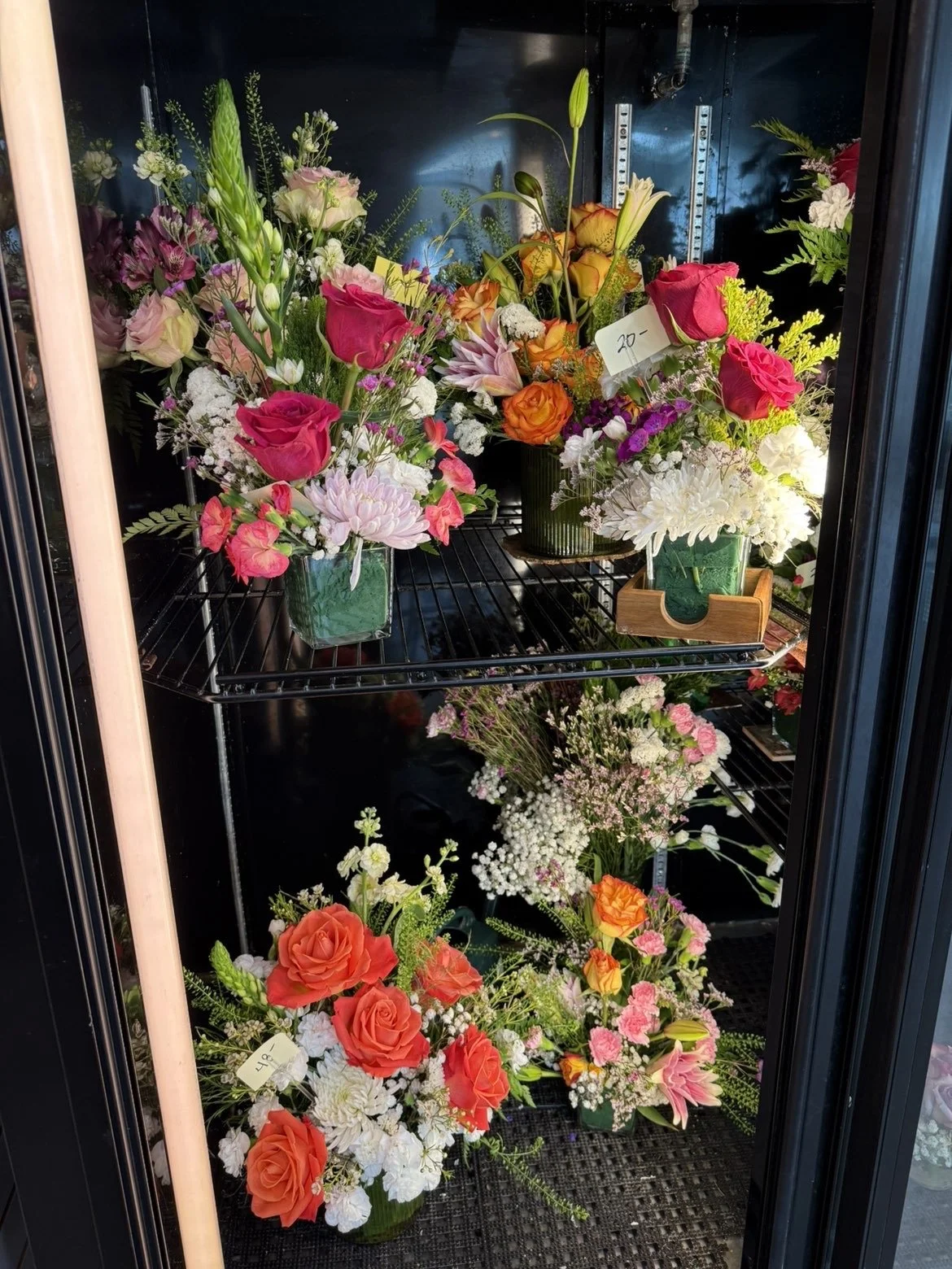 Vitrine de fleurs fraîches avec diverses bouquets colorés, comprenant des roses rose et orange, des chrysanthèmes blancs, des lys, et d'autres fleurs dans différents contenants.