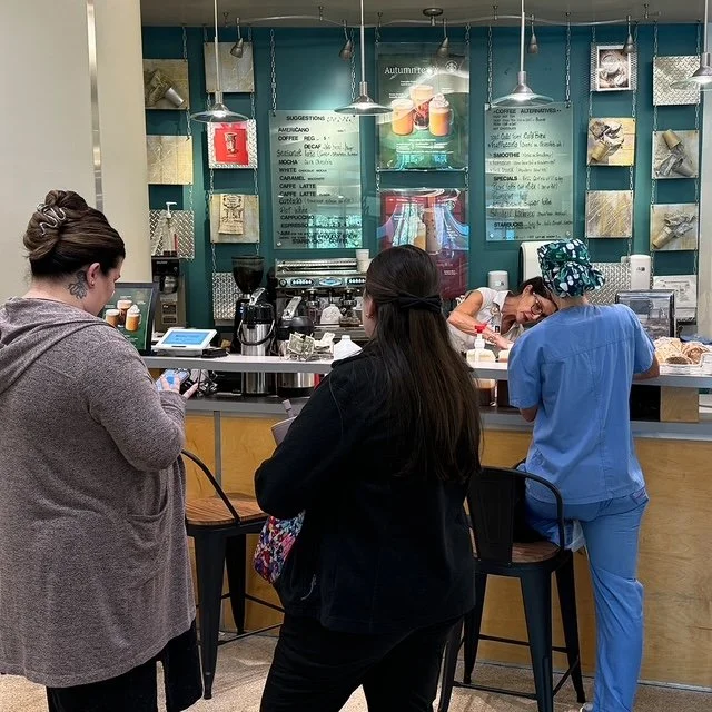 Trois personnes dans un café, un employé derrière le comptoir prépare une commande, deux clientes attendent, une parlant à son téléphone, l'autre regarde le comptoir.