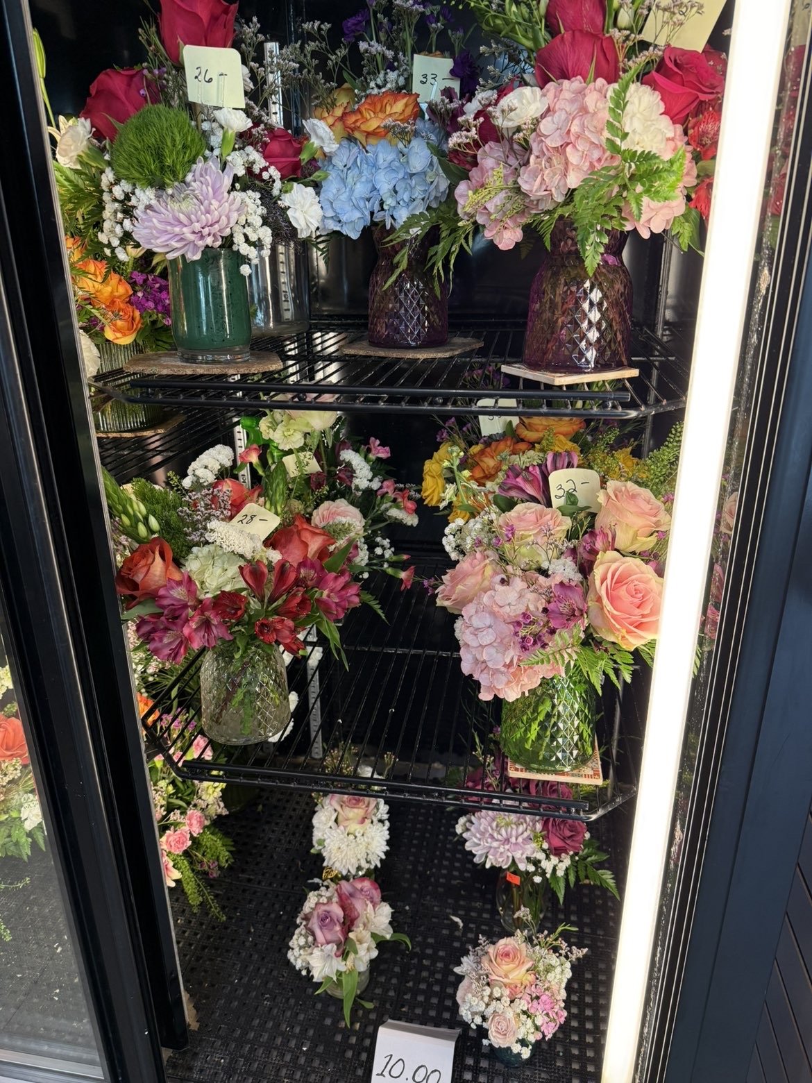 Vitrine réfrigérée contenant diverses compositions florales dans des vases en verre colorés avec des étiquettes de prix.