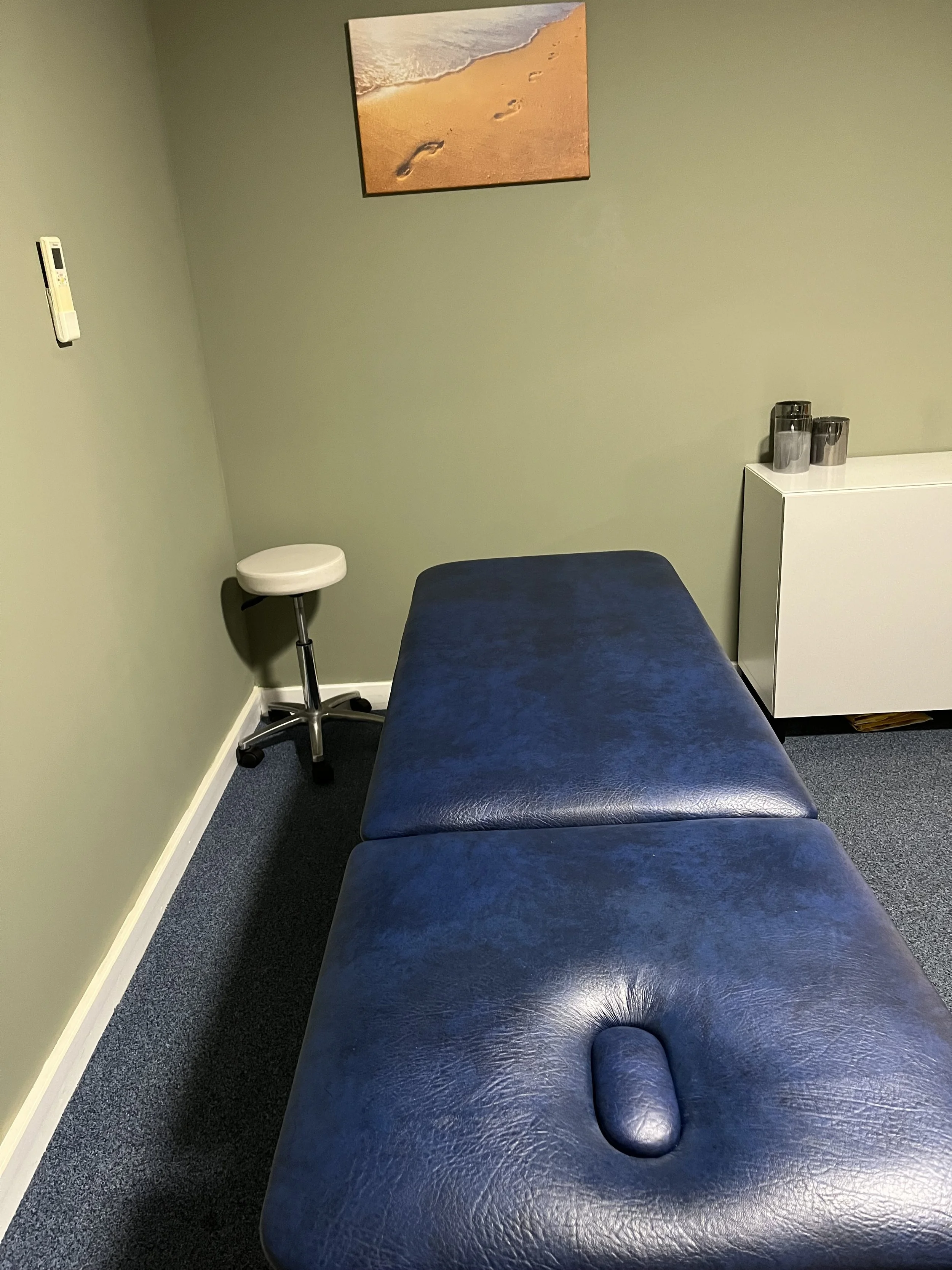 Blue padded examination table in a small medical treatment room with a rolling stool, cabinet, supplies, and beach artwork.