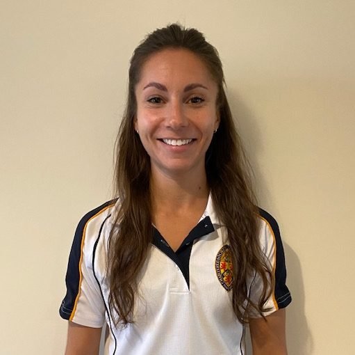 A young woman with long brown hair, smiling, wearing a white sports polo shirt with black and gold accents and an emblem on the left chest.