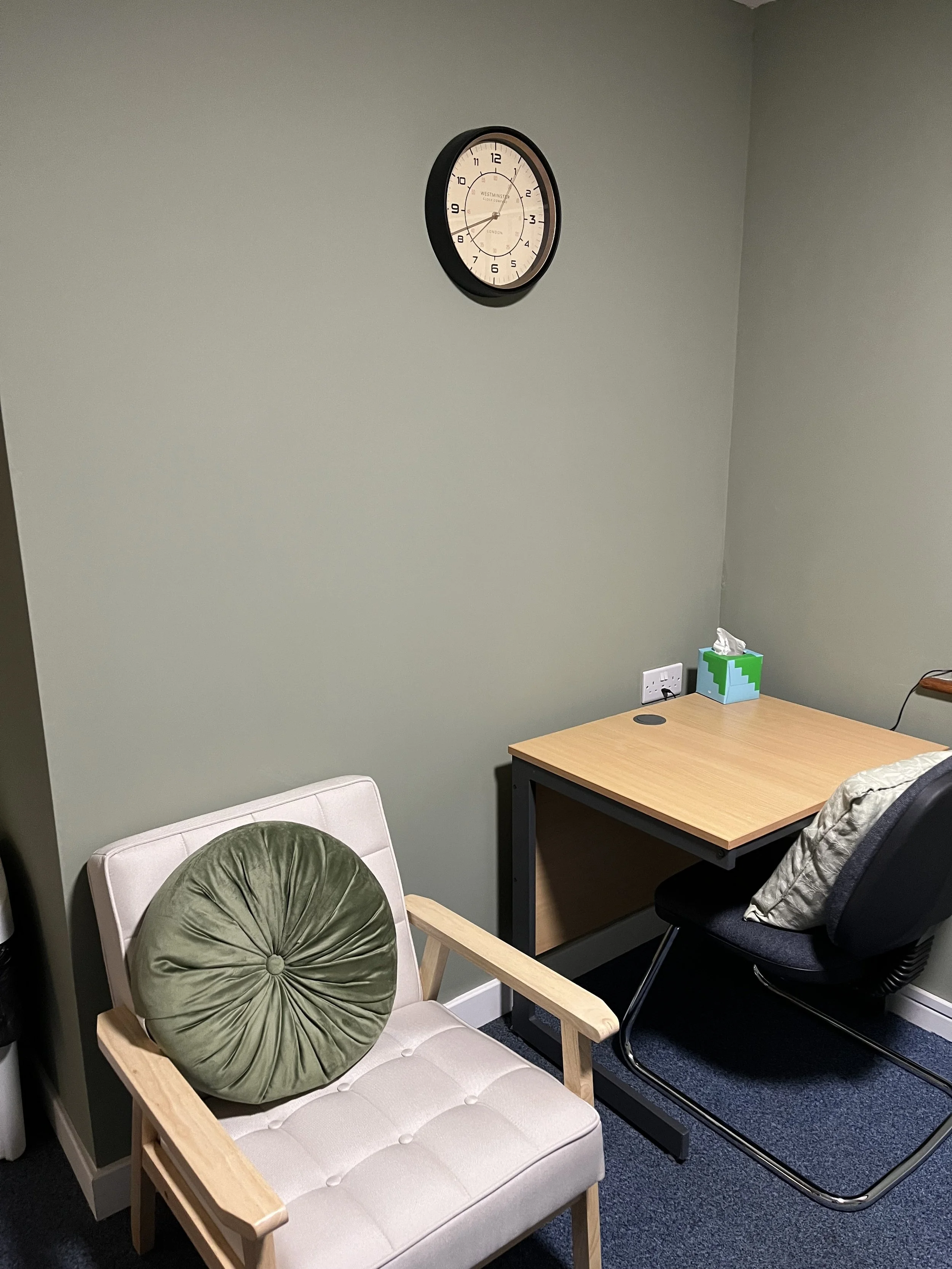 Home office with a wall clock, wooden desk, tissue box, office chair, and cushioned armchair