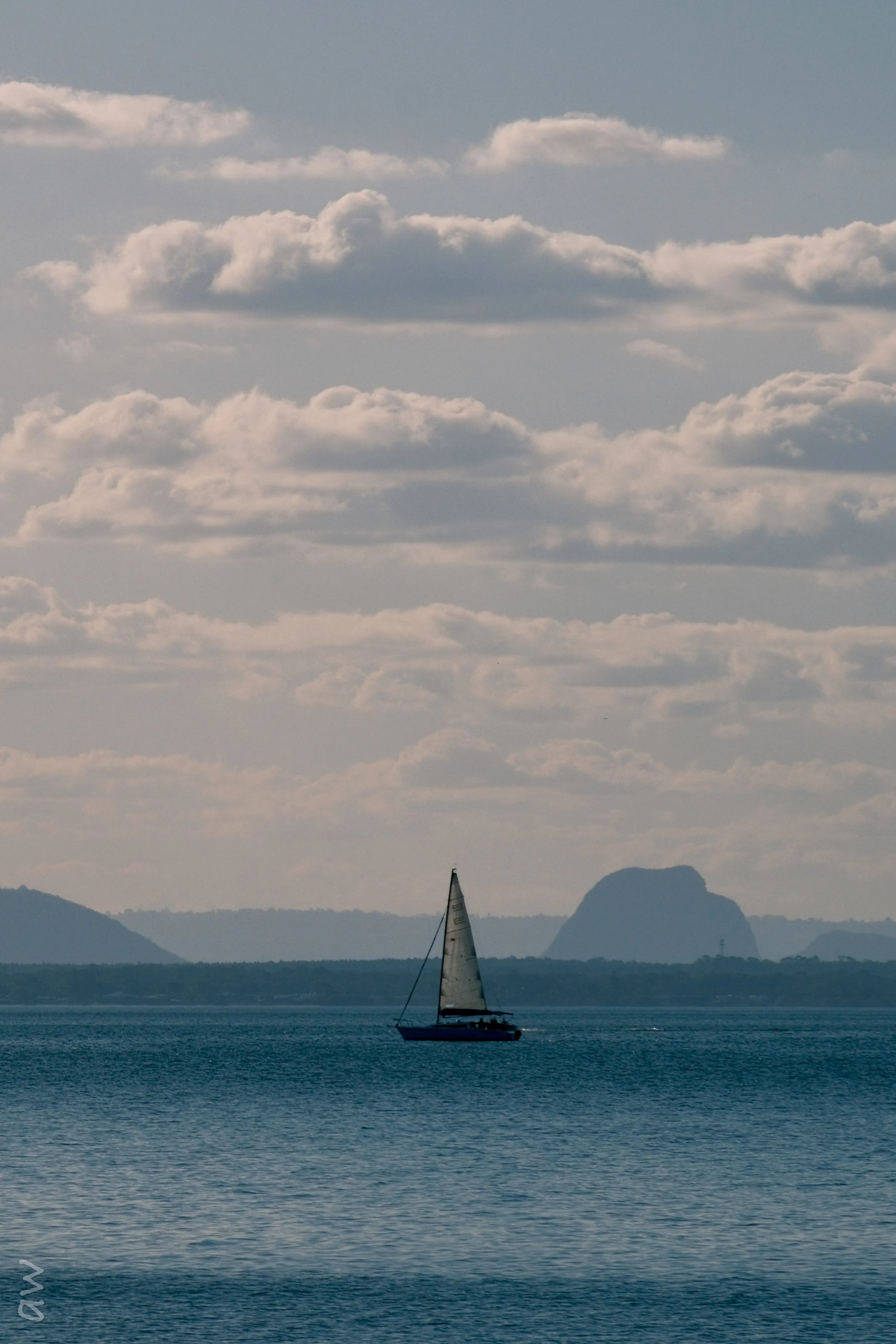 A sailboat beneath a cloud-filled sky, with distant mountains across the water.