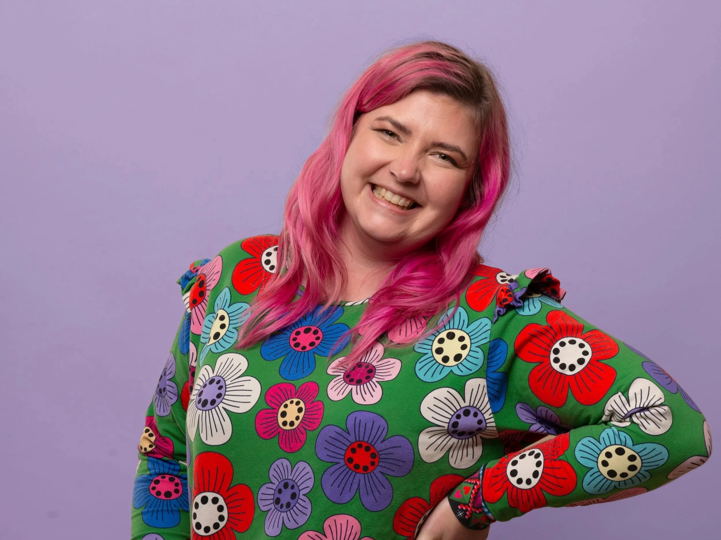 A woman with pink hair, smiling, wearing a colorful, floral-patterned shirt, standing against a light purple background.