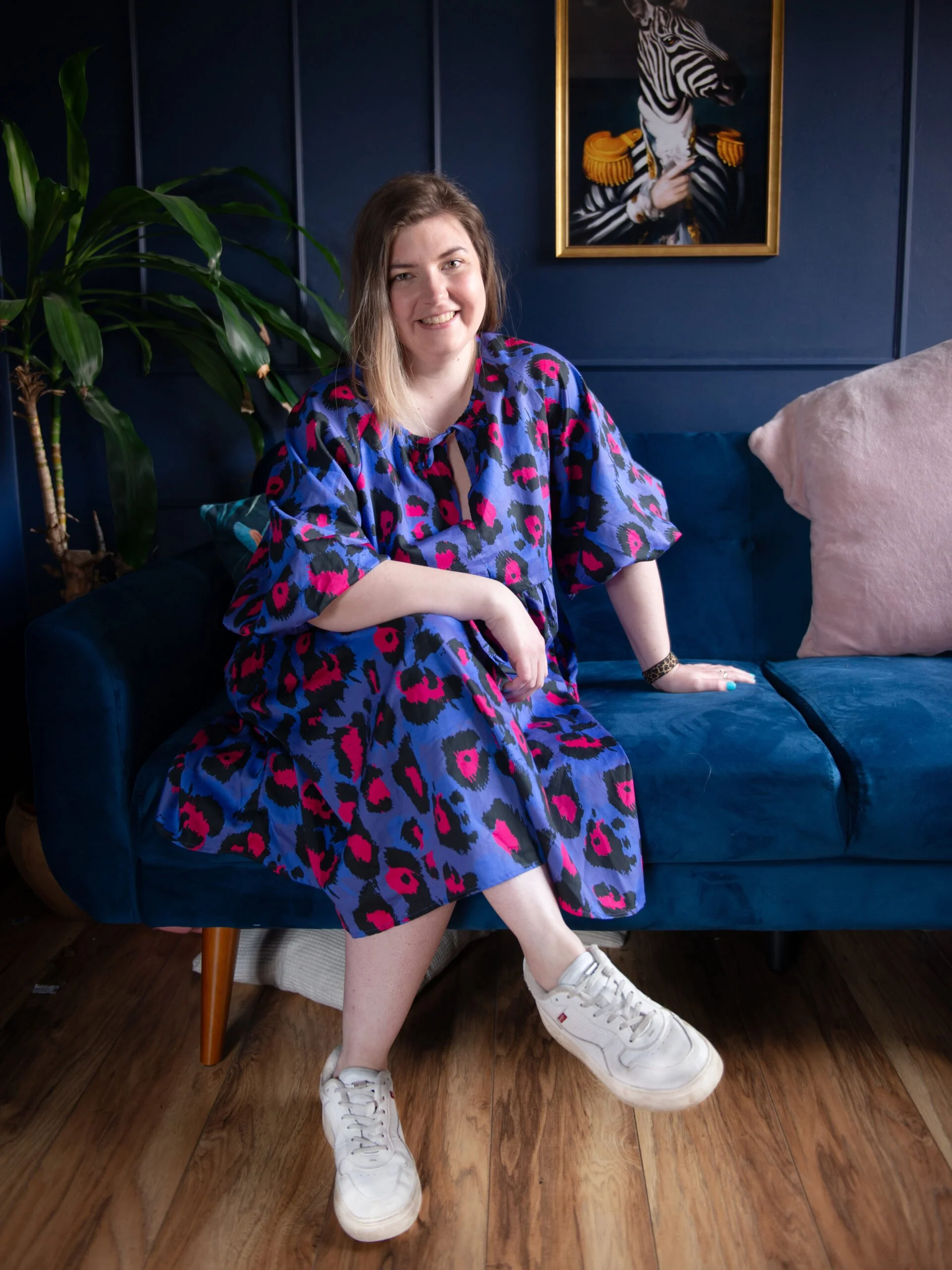 A woman with shoulder-length hair sitting on a blue velvet couch, smiling, wearing a colorful leopard print dress, white sneakers, and a bracelet, with a large indoor plant and a framed zebra portrait on a dark blue wall behind her, and a beige cushion on the couch.