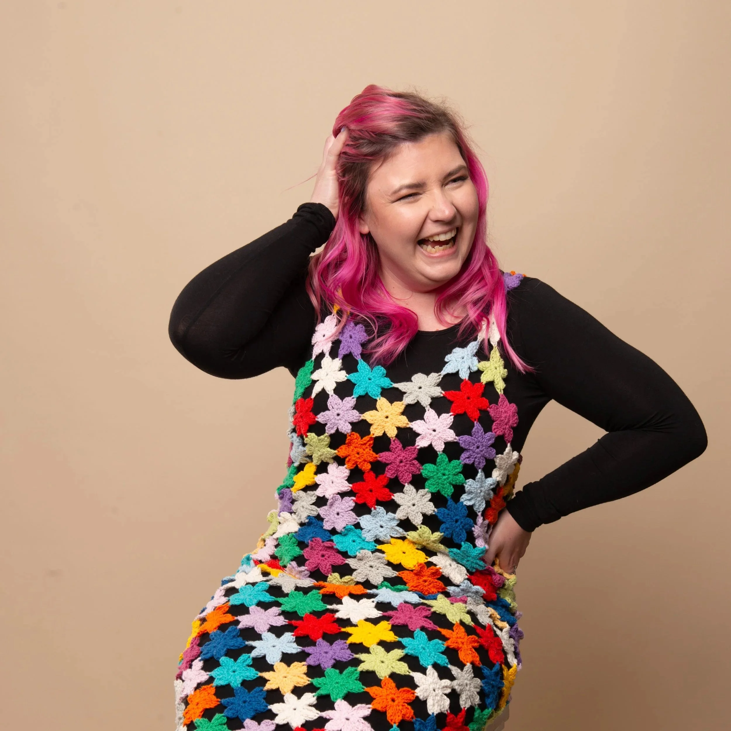 A woman with pink hair wearing a black shirt with colorful crocheted flowers. She is smiling and touching her head with her right hand, standing against a beige wall.