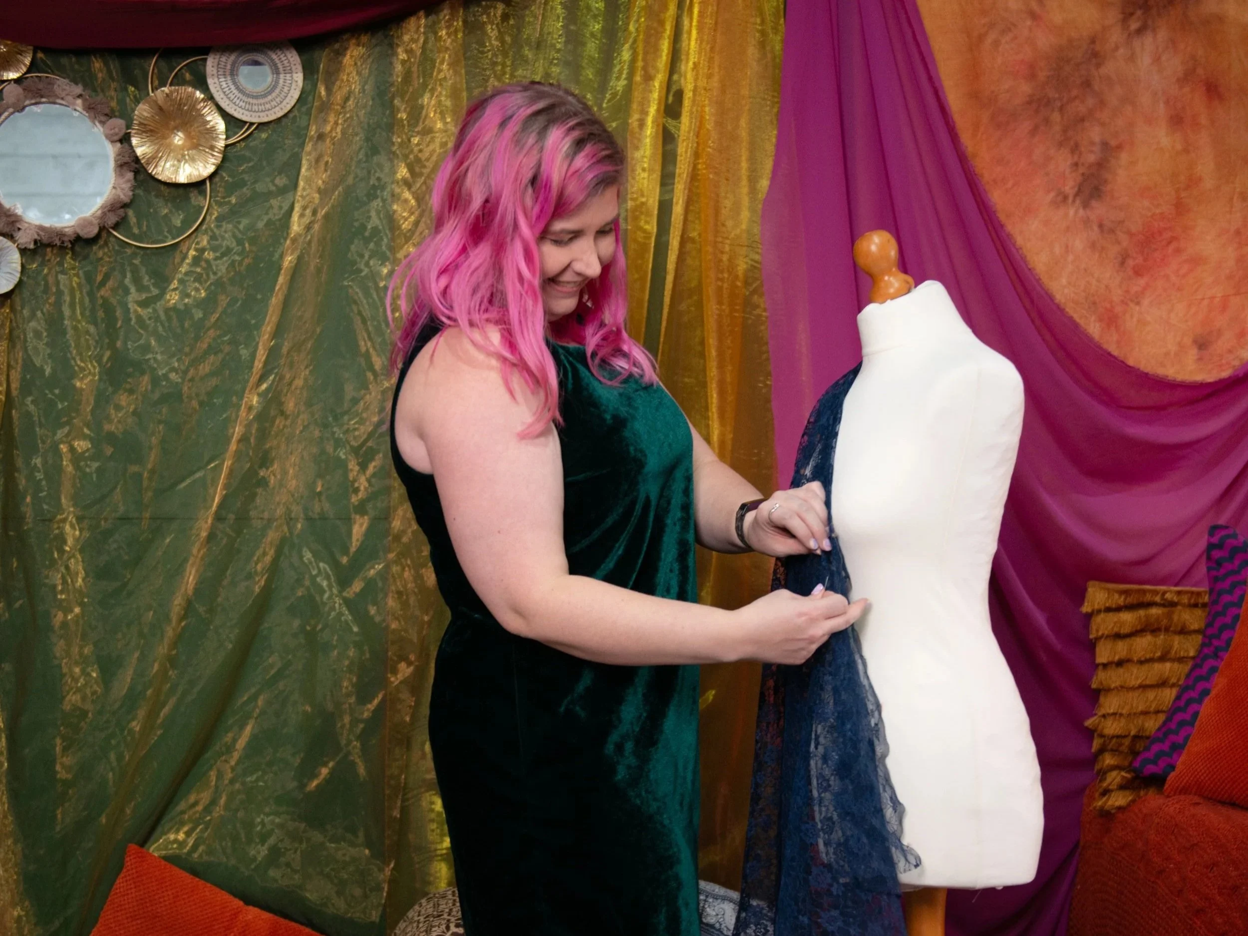 A woman with pink hair smiling as she looks at a dress form, with a vibrant, colorful background of draped fabric and decorative mirrors.