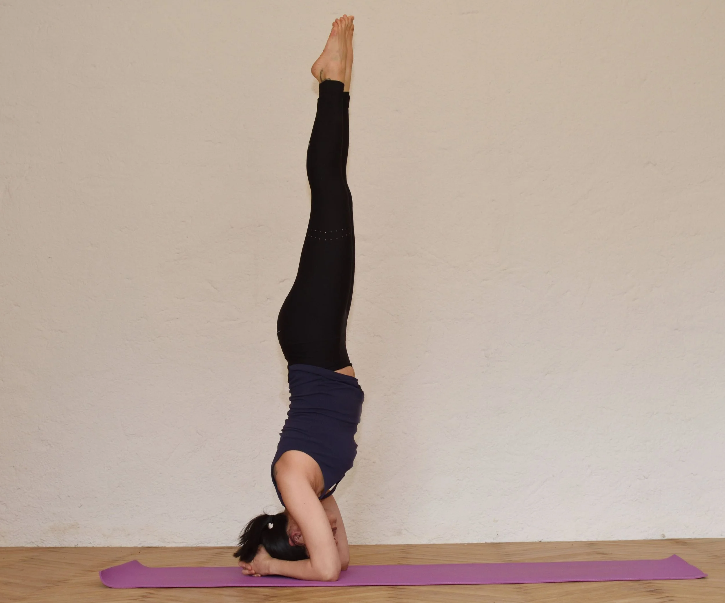 A woman performing a headstand yoga pose on a pink yoga mat against a plain wall.
