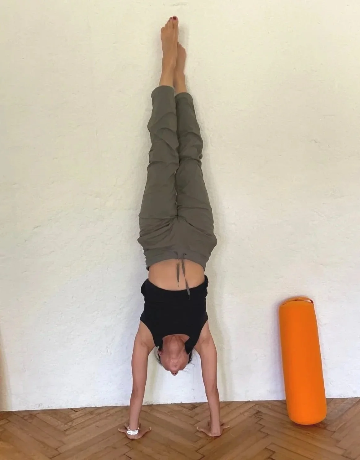 Person performing a handstand against a white wall, with their legs extended vertical and feet touching the wall. An orange yoga bolster is beside the person.