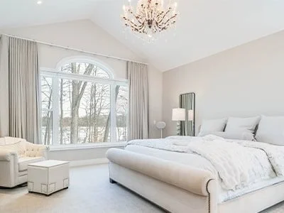 Bright bedroom with large window, white bed, chandelier, and soft furnishings.