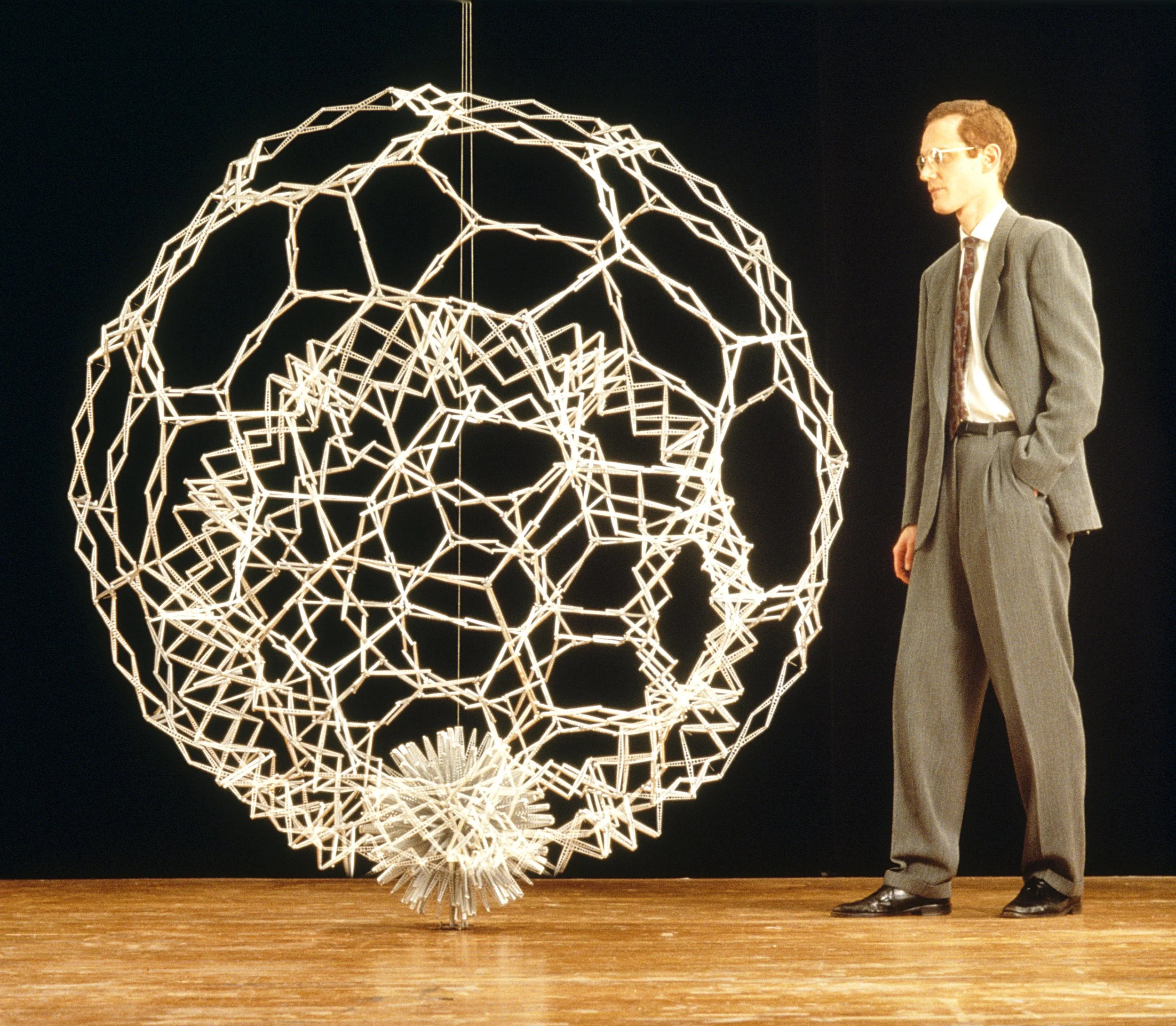 A man in a gray suit and glasses stands next to a large illuminated wire sculpture of a human head. The sculpture is made of interconnected geometric shapes and glowing lights, positioned against a dark background. The man is looking at the sculpture with his hands in his pockets.