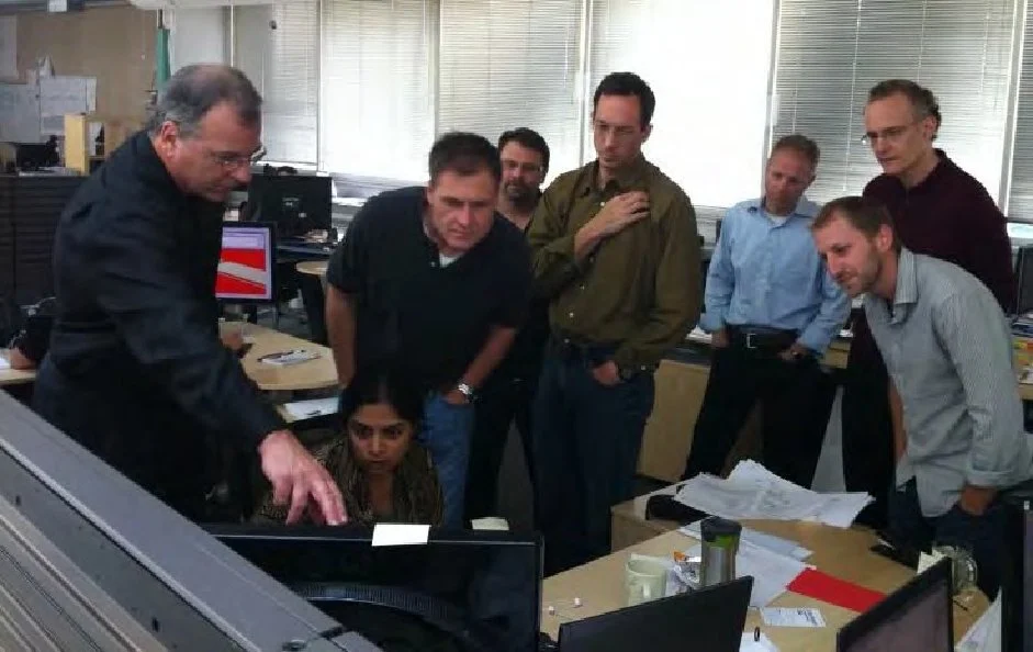 Group of seven professionals gathered around a computer in an office, looking at the screen with serious expressions.