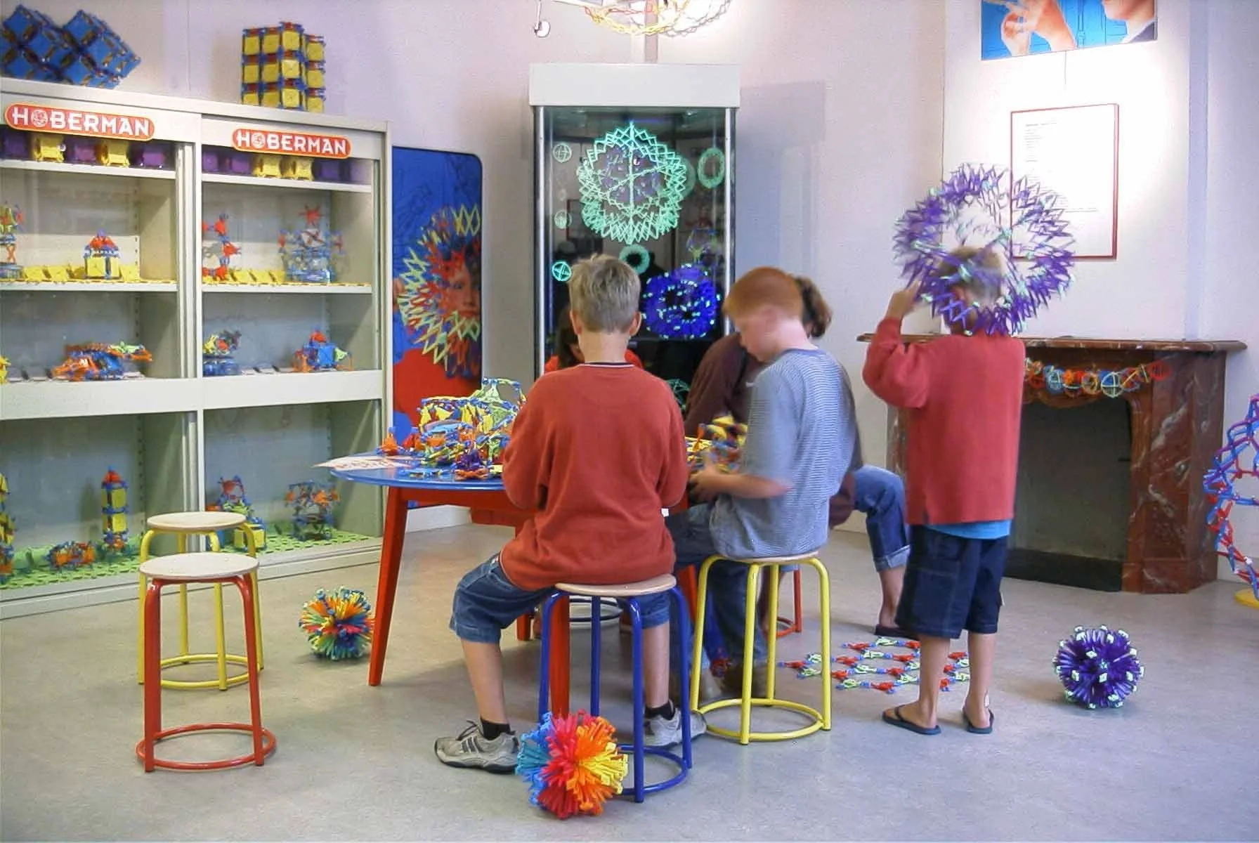Children assembling colorful geometric models in a classroom filled with display cases and artwork.