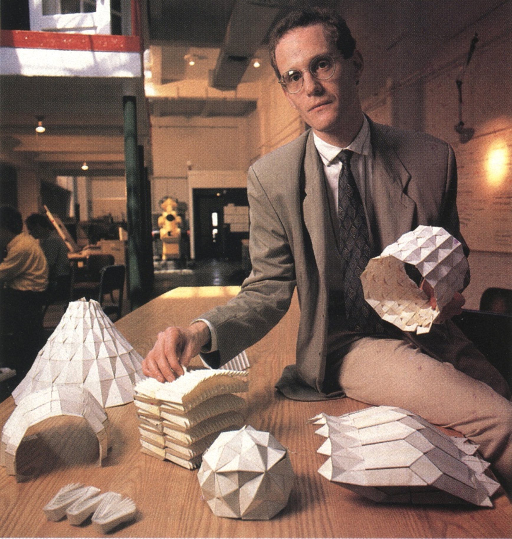 A man in a gray suit and glasses is sitting at a table in a room, shaking hands with a person outside the frame. On the table are multiple geometric, faceted sculptures of owls made from a white, textured material, some stacked. In the background, there are people sitting at tables, a yellow flower arrangement, and warm lighting.