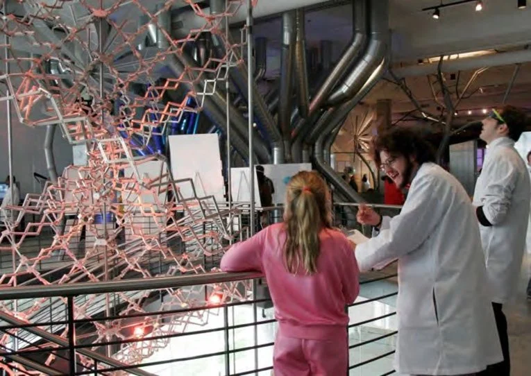 A woman in a white lab coat and glasses talking to a young girl with long blond hair, wearing a pink sweater, in a science or technology exhibit. The background features an intricate metallic structure and large industrial pipes.