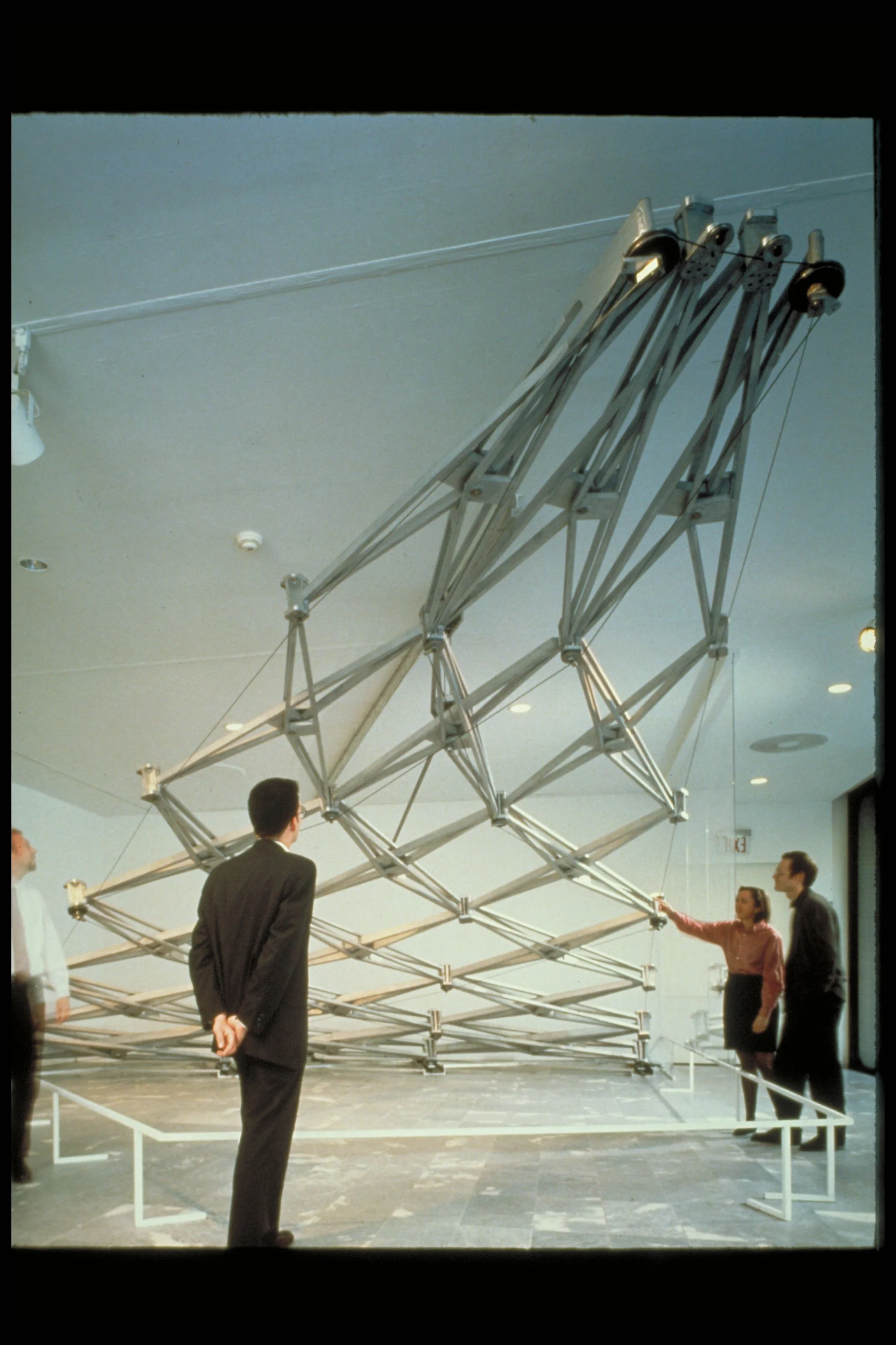 Museum exhibit of a large metallic sculpture resembling a geometric crab with multiple legs, with several people observing it.