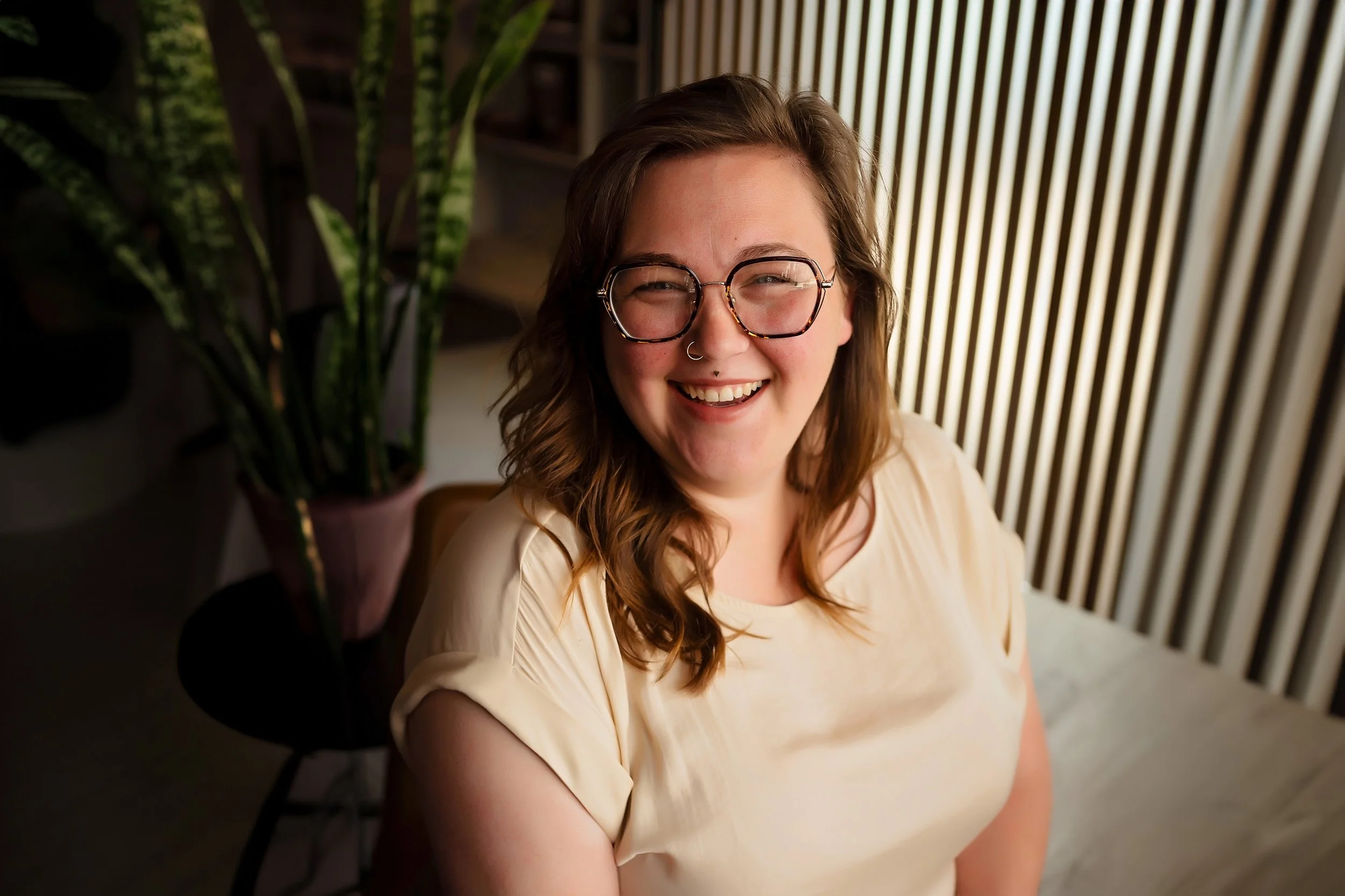 Kirsten Lane with long, wavy brown hair, wearing glasses and a septum piercing, is smiling and sitting indoors near a window with vertical blinds. A potted plant is visible on the left side of the image.