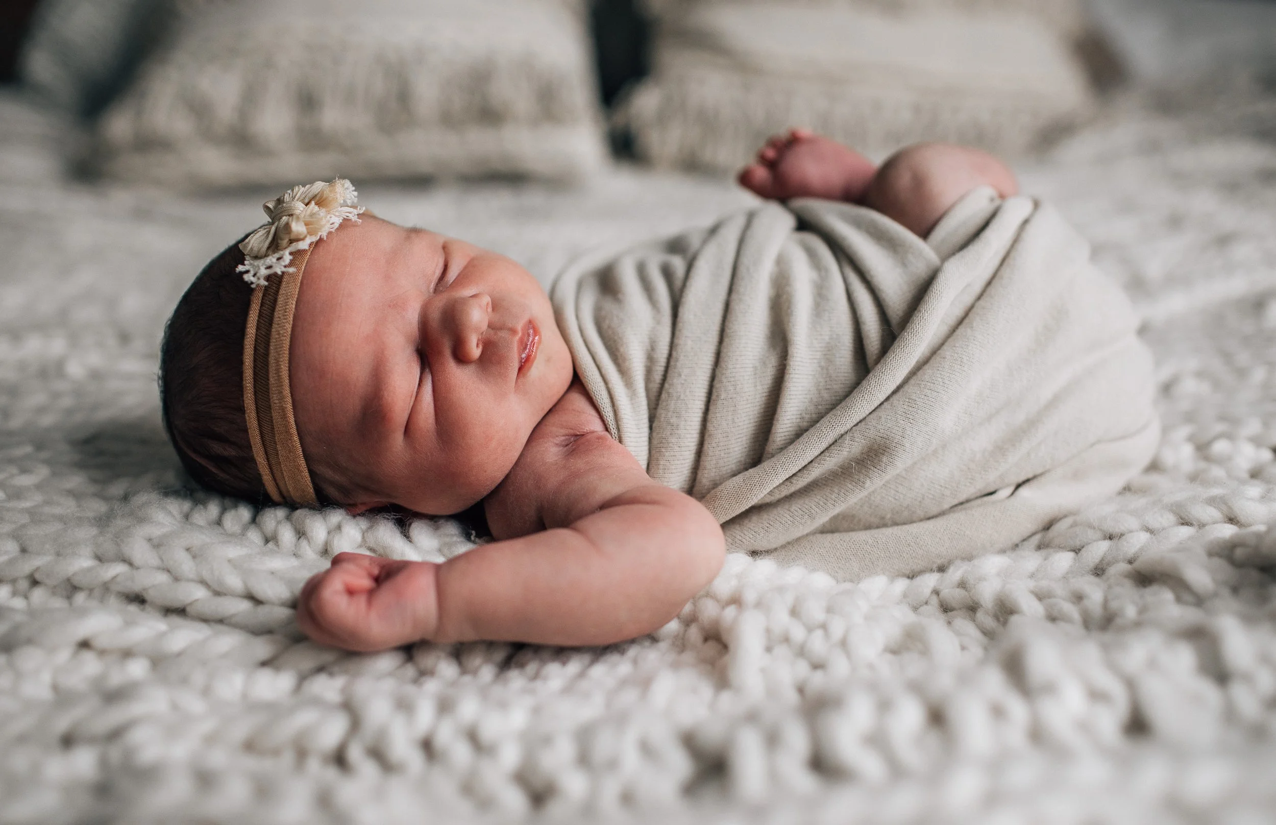 A sleeping newborn baby lying on a soft, knitted blanket. The baby is wrapped in a light-colored fabric and is wearing a delicate headband with a small bow.