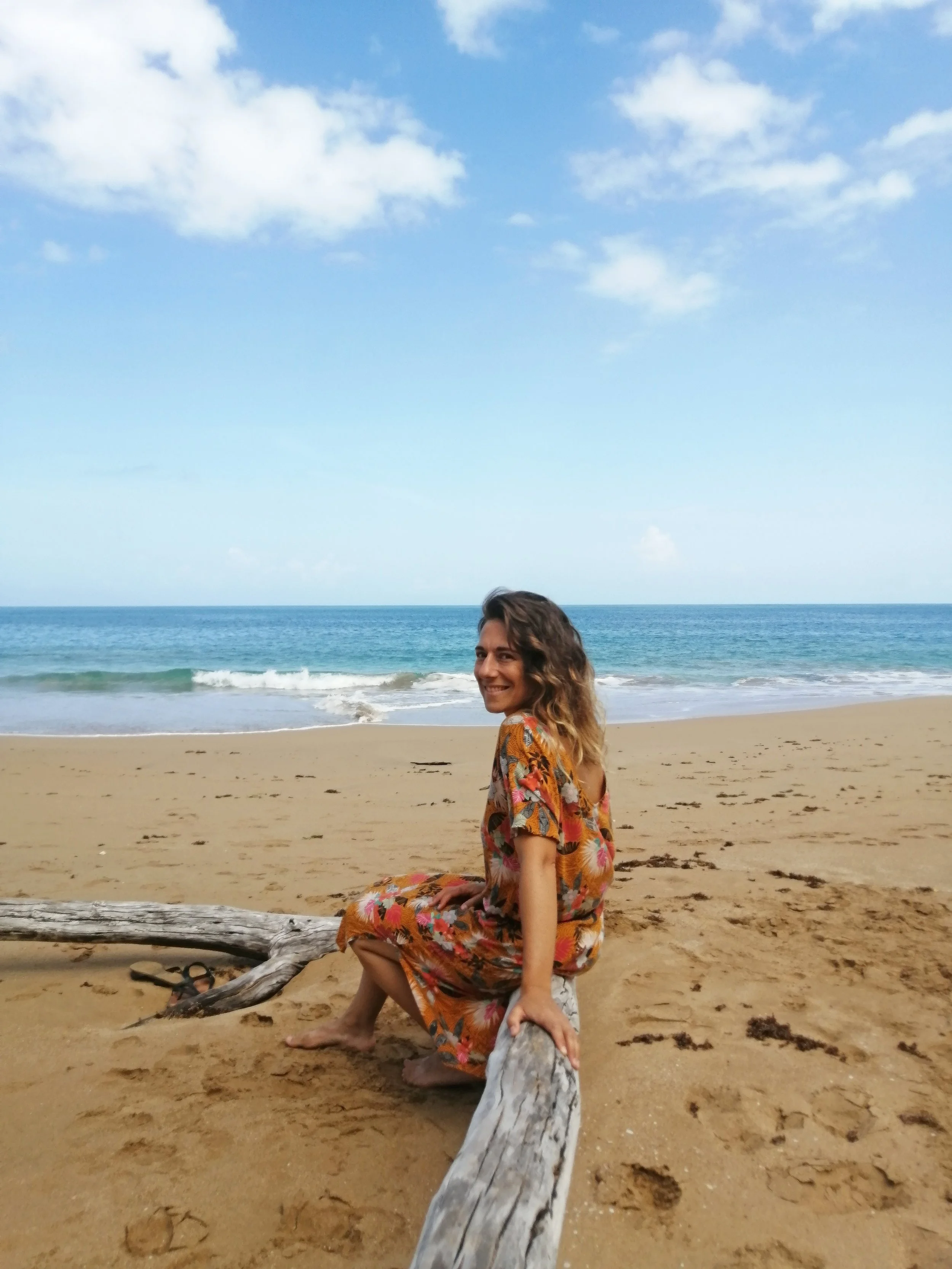 Femme assise sur la plage avec le sourire, portant une robe à motifs colorés, près d'une branche d'arbre couché sur le sable, arrière-plan de mer calme et ciel bleu avec quelques nuages.