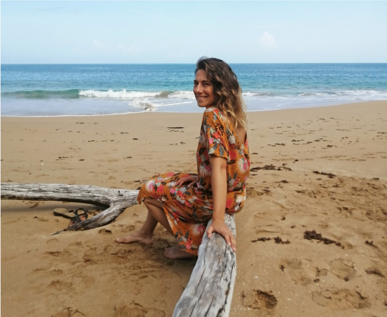 Une femme souriante assise sur un bois flotté sur une plage de sable, avec la mer en arrière-plan.