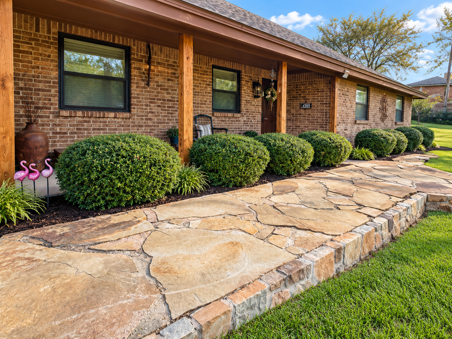 A brick house with a front porch supported by wooden posts. The porch has a bench, a wall-mounted lantern, and a decorative wreath on the door. In front of the porch, there is a stone pathway bordered by well-maintained bushes and small plants. A large decorative vase and pink flamingo statues are also visible near the bushes. The surrounding area includes a grassy lawn and a background of trees under a blue sky with clouds.