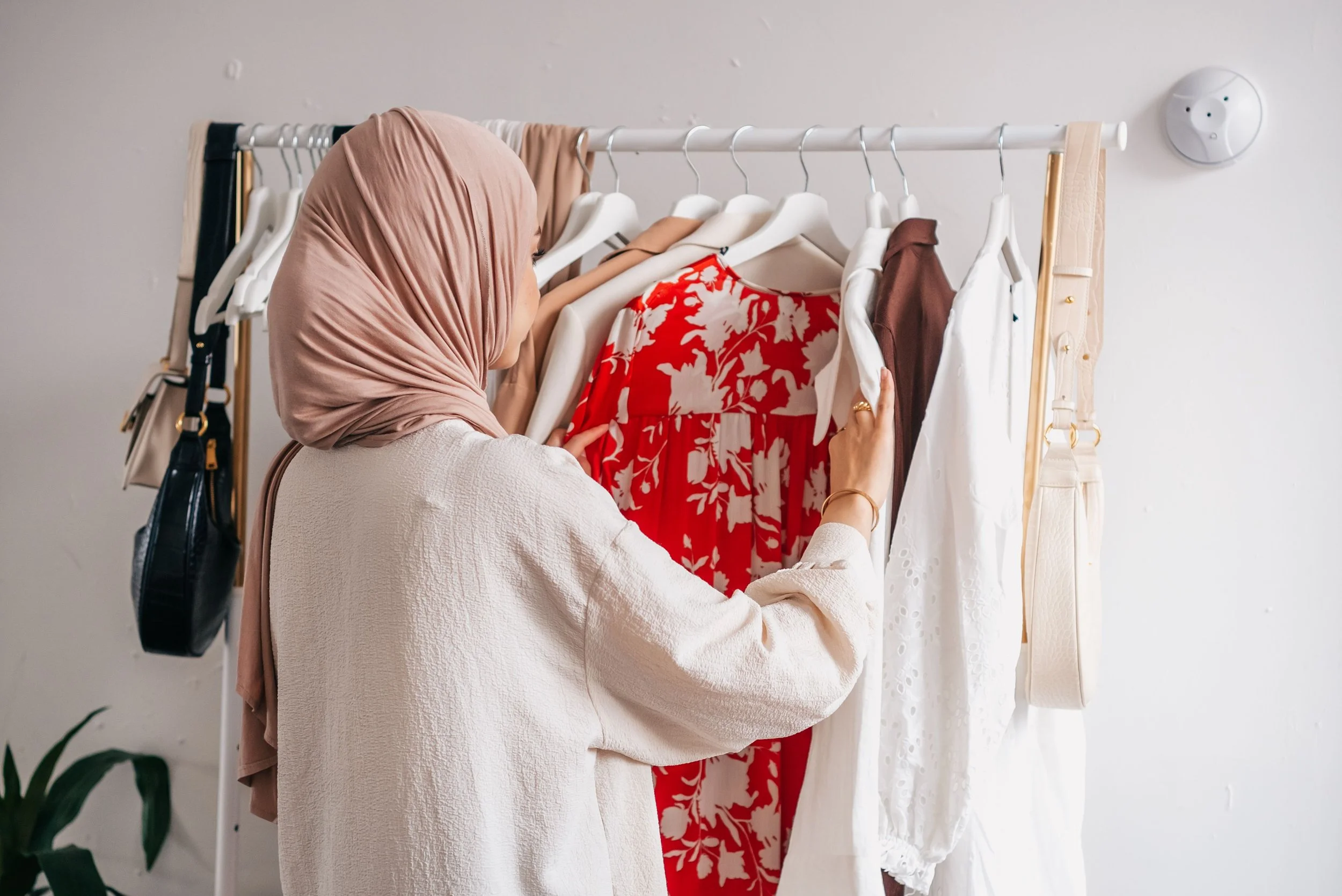 A woman wearing a light pink hijab and a white textured top is looking at clothes hanging on a white clothing rack. The rack has various dresses and garments, including a red and white floral dress, a brown dress, and white dresses with eyelet details. The background shows a plain wall with a circular device or thermostat, and part of a plant is visible in the lower left corner.