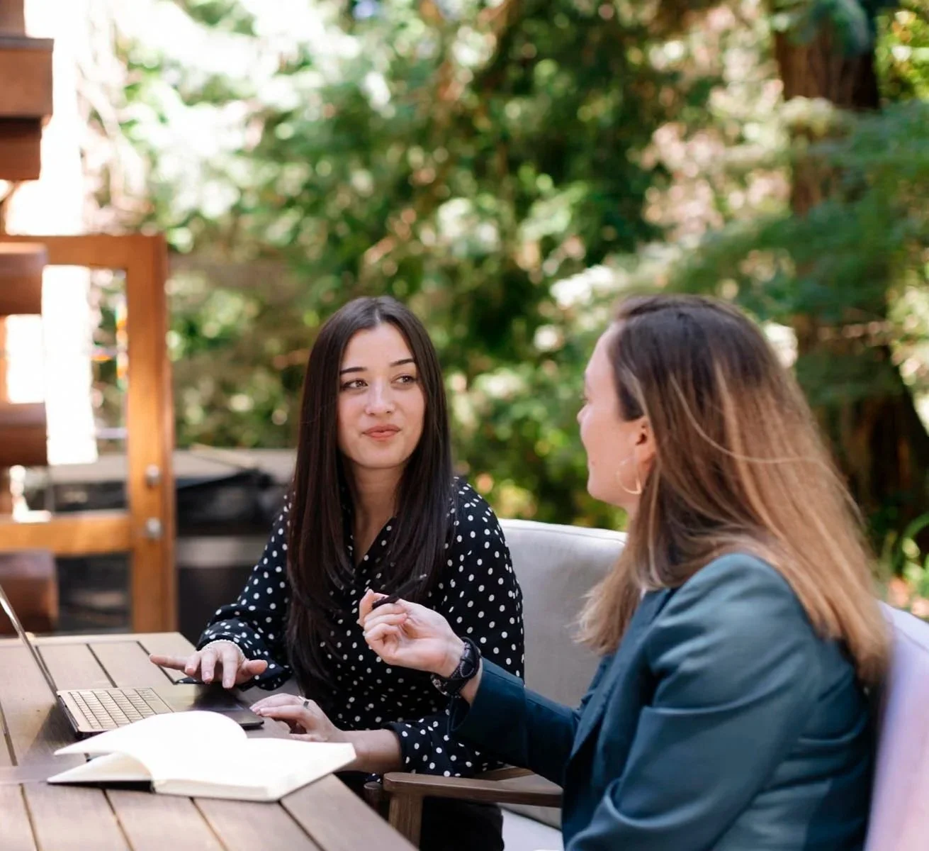 Two women having a conversation outdoors at a table with a laptop, notebook, and pen, surrounded by greenery.