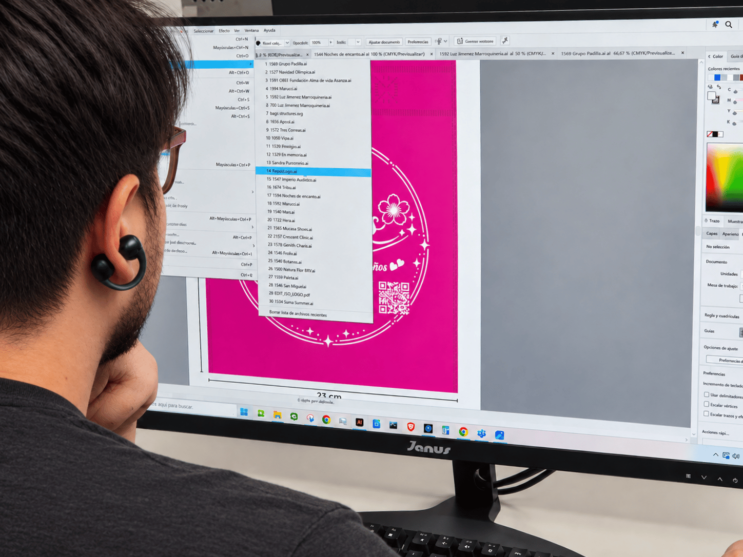 A person working on a large computer screen, designing a pink graphic with white decorative elements and a QR code. The screen displays a design software interface with various menus and tools.