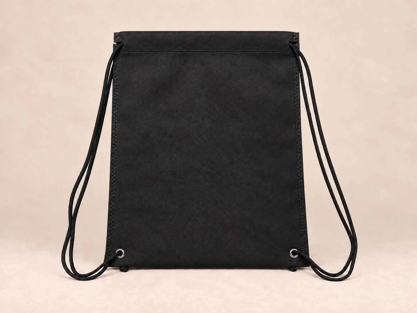 Black drawstring backpack made of fabric with metal eyelets and black cords on a plain beige background.