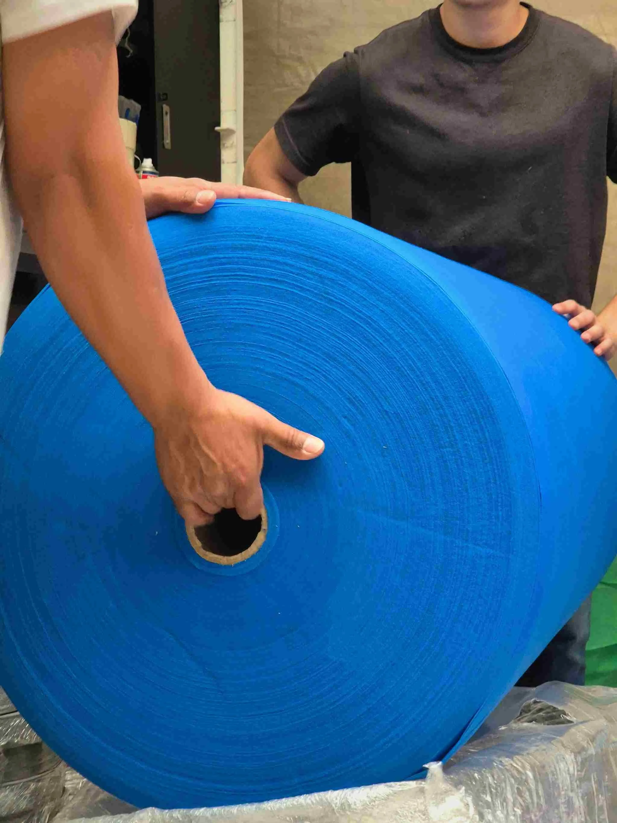 A person holding a large roll of material cambrel, with another person partially visible in the background, in an indoor setting.