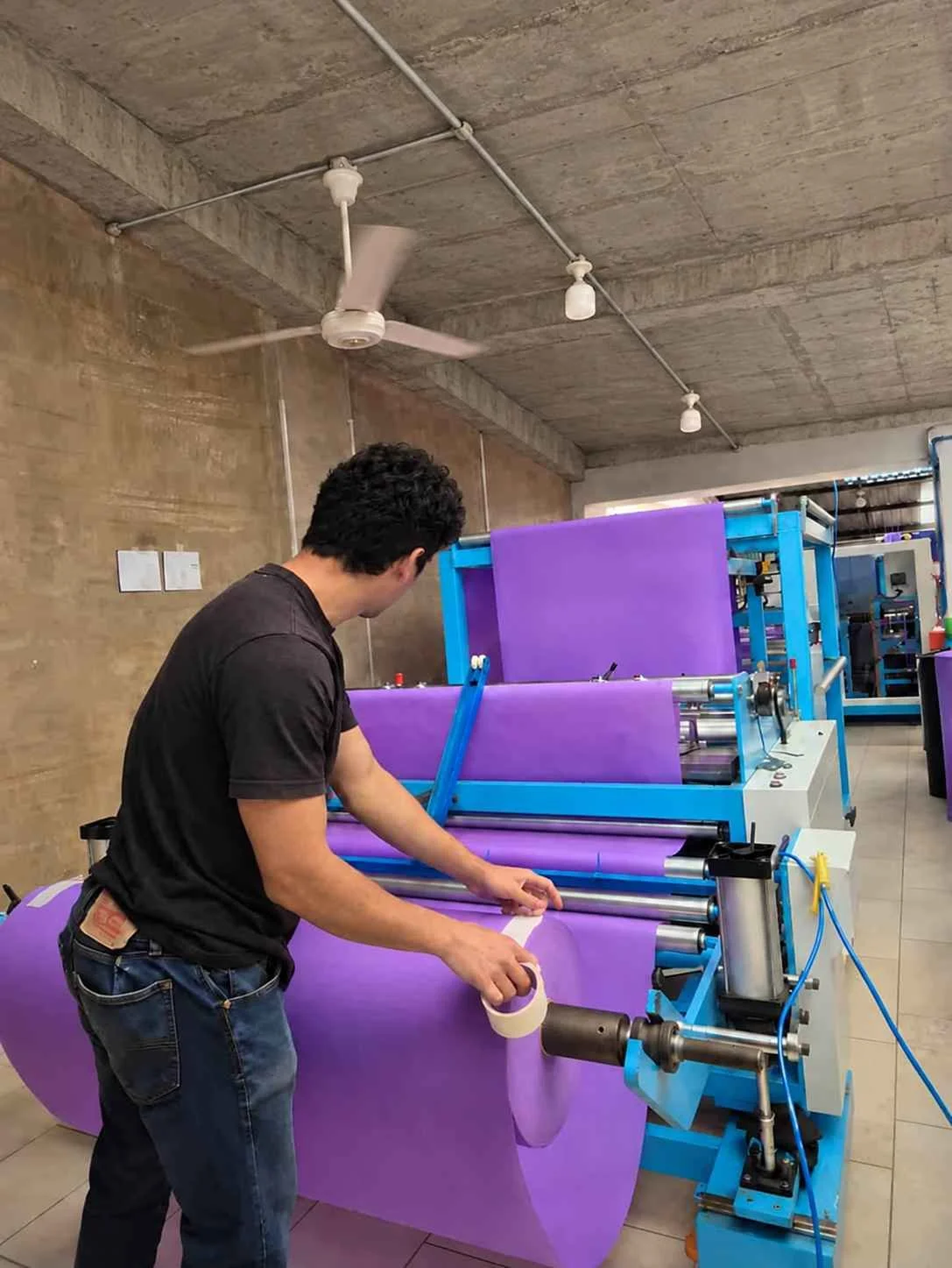 A man working with a large machine ultrasonido de fabricación de bolsas, handling a roll of purple material in an industrial setting with concrete walls and ceiling.