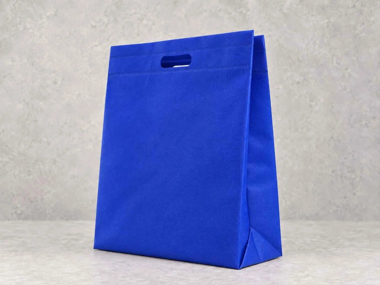 A blue cambrel shopping bag with a cut-out handle on a plain light-colored background.
