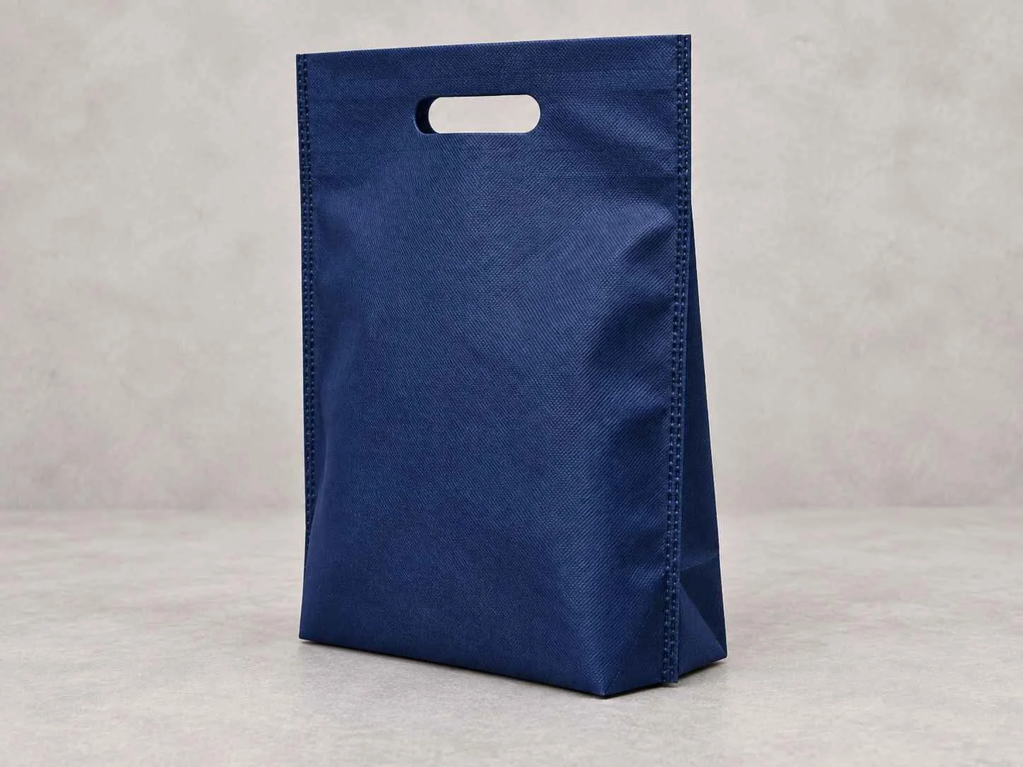 	
Una bolsa reutilizable azul oscuro de cambrel plana troquelada con fuelle abajo de pie vertical sobre una superficie neutra contra un fondo neutro.
