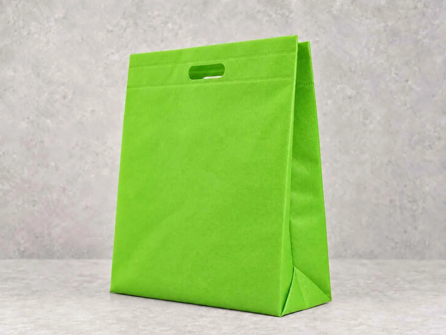 A bright green reusable shopping bag standing upright on a light gray surface with a textured gray wall in the background. Bolsa en cambre con troquel 3 fuelles. Color verde