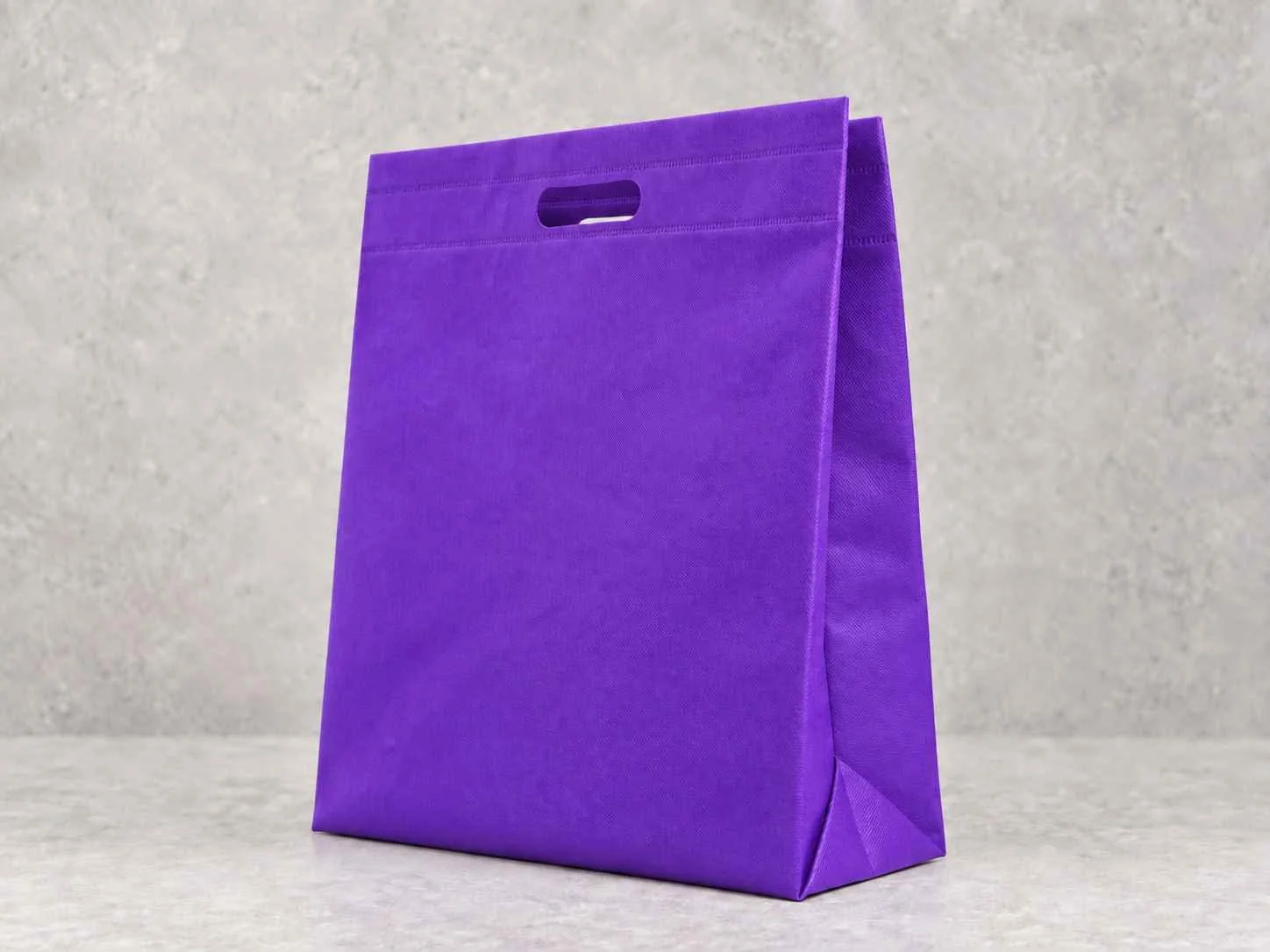 A purple reusable shopping bag standing upright on a light-colored surface against a light gray background. Bolsa en cambre con troquel 3 fuelles. Color morada
