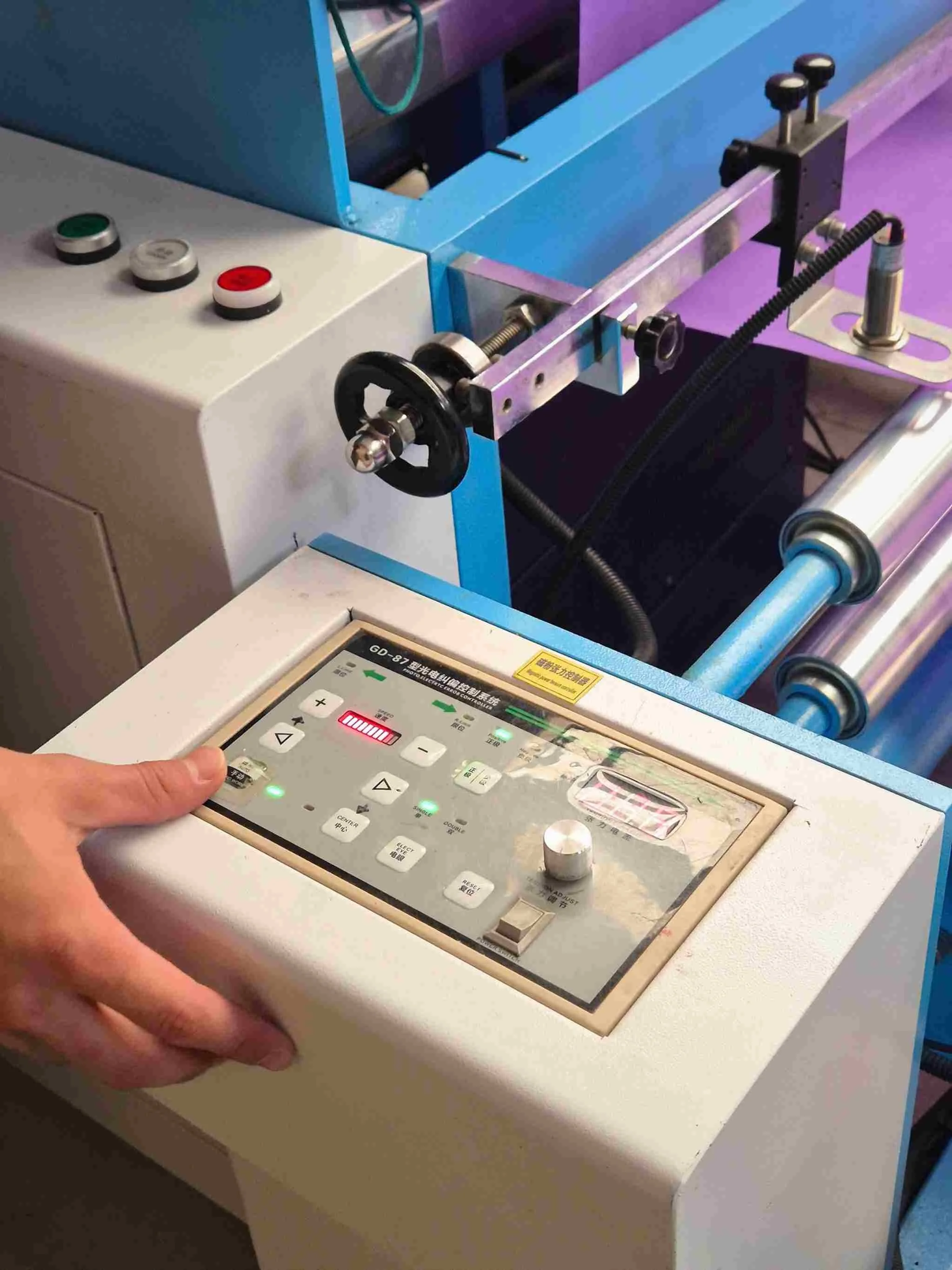 Close-up of an electronic control panel with various buttons and a hand pressing a button, adjacent to a machine part with rollers and metal components.