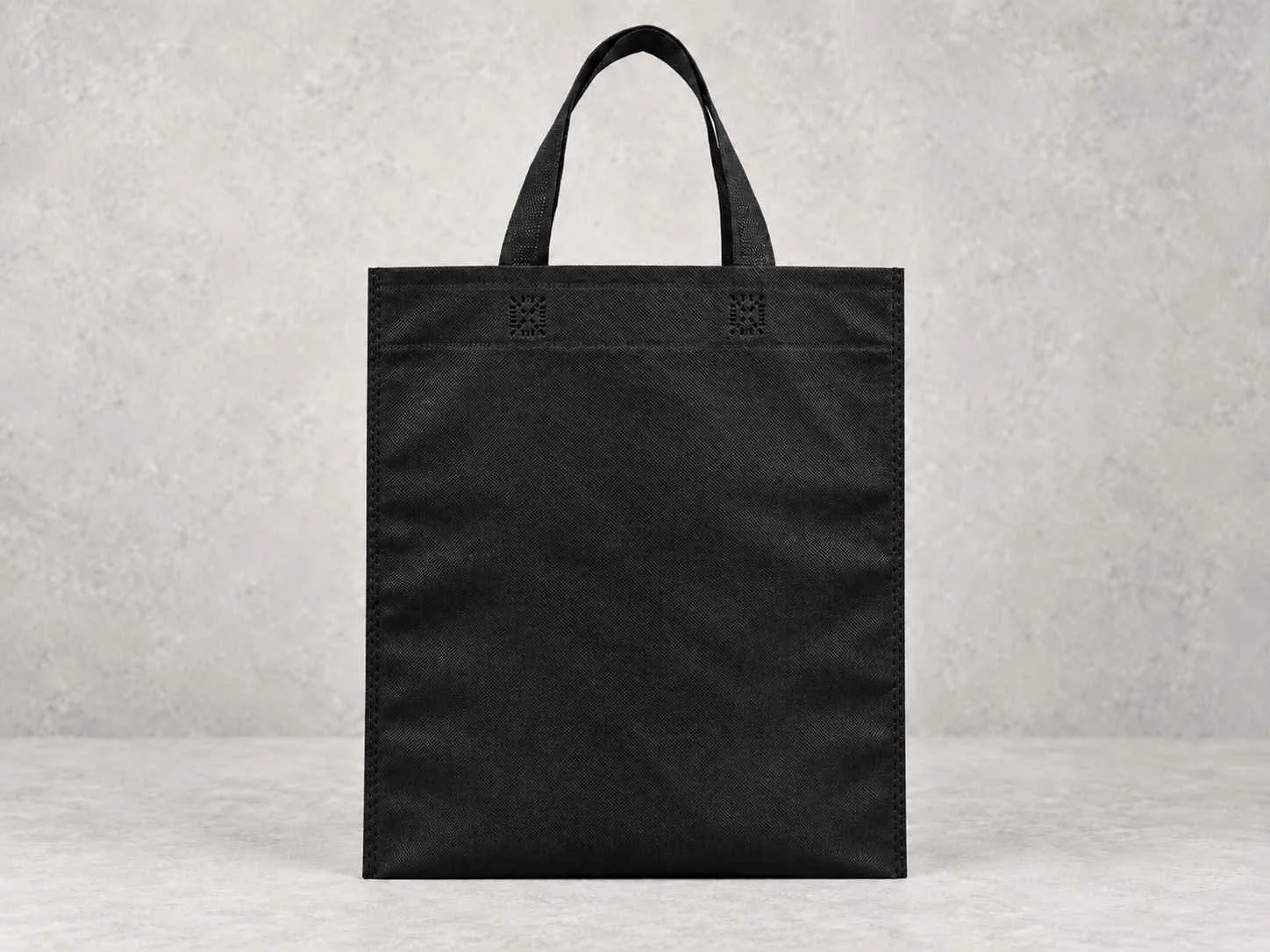 A black reusable tote bag with small handles, placed against a neutral gray background.