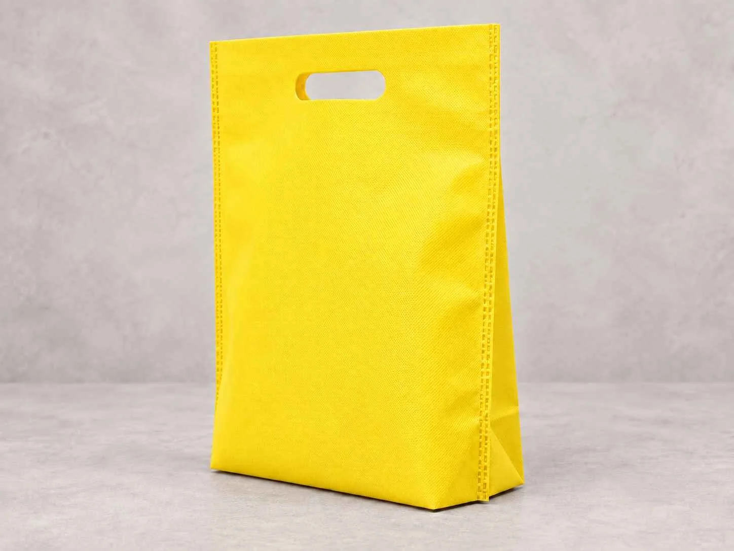 	
Una bolsa reutilizable amarilla de cambrel plana troquelada con fuelle abajo de pie vertical sobre una superficie neutra contra un fondo neutro.