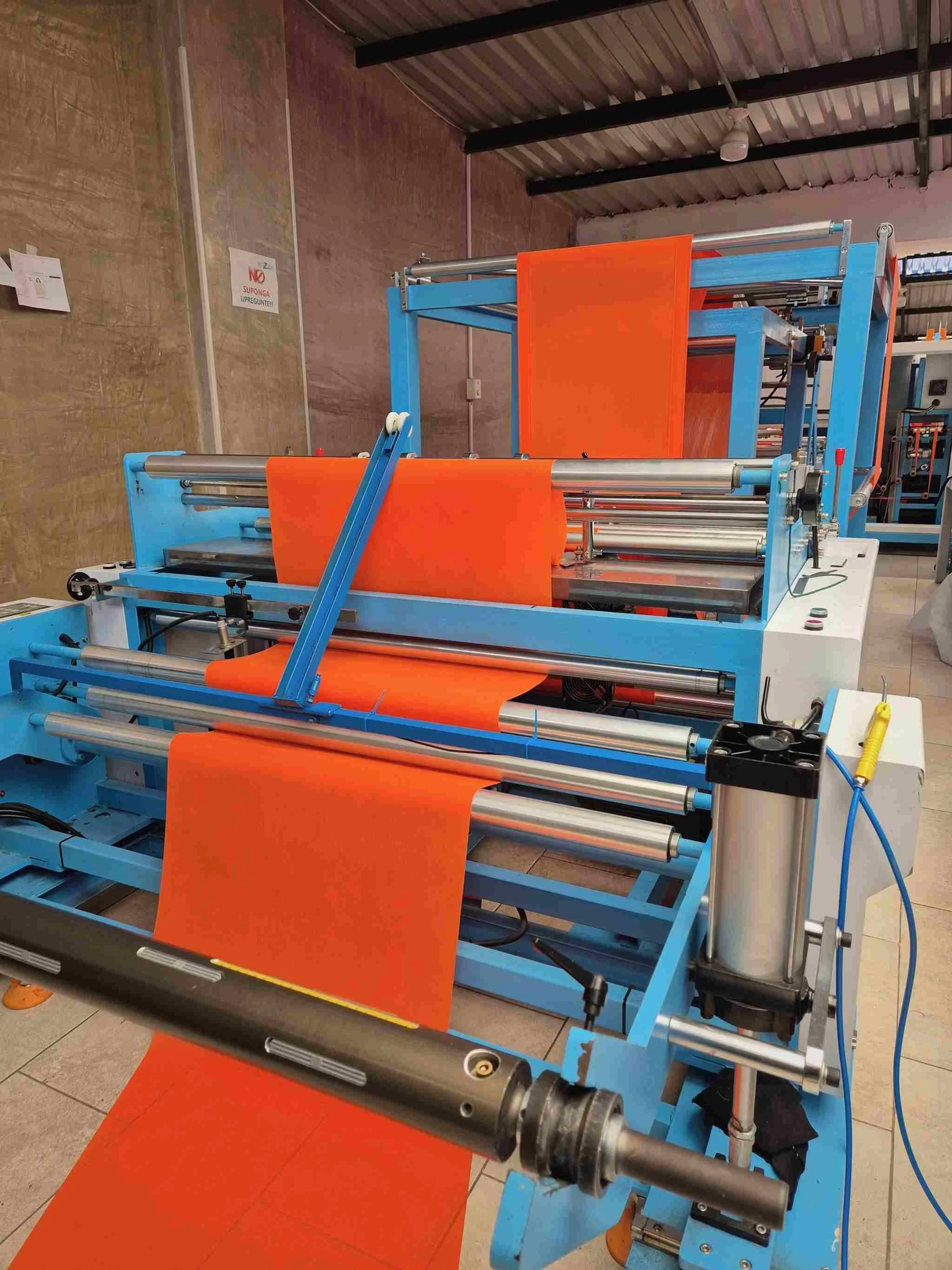 A machine ultrasonido producing tela cambre orange, possibly in a manufacturing or industrial setting with a metal roof and tiled floor.