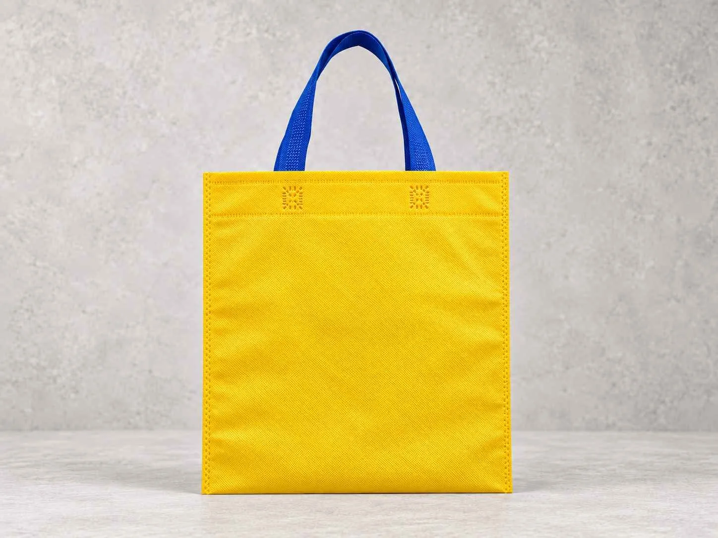 Yellow fabric shopping bag with blue handles on a light-colored surface against a neutral background.