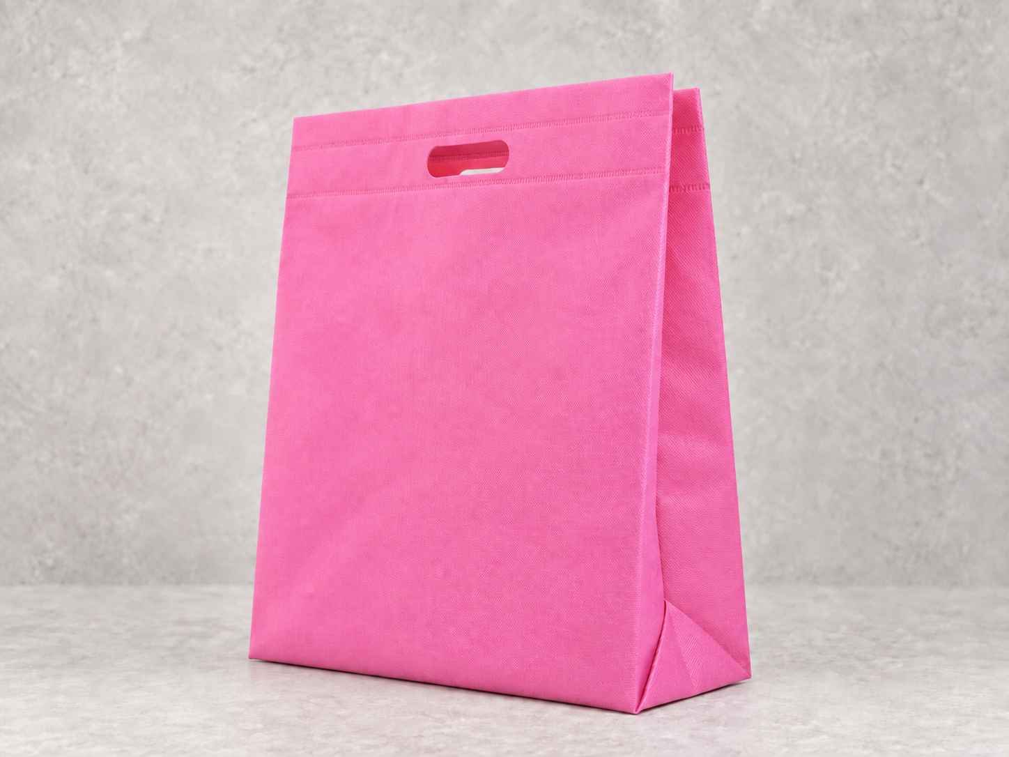 Pink reusable shopping cambrel bag with cutout handles, standing on a light-colored surface against a gray wall.