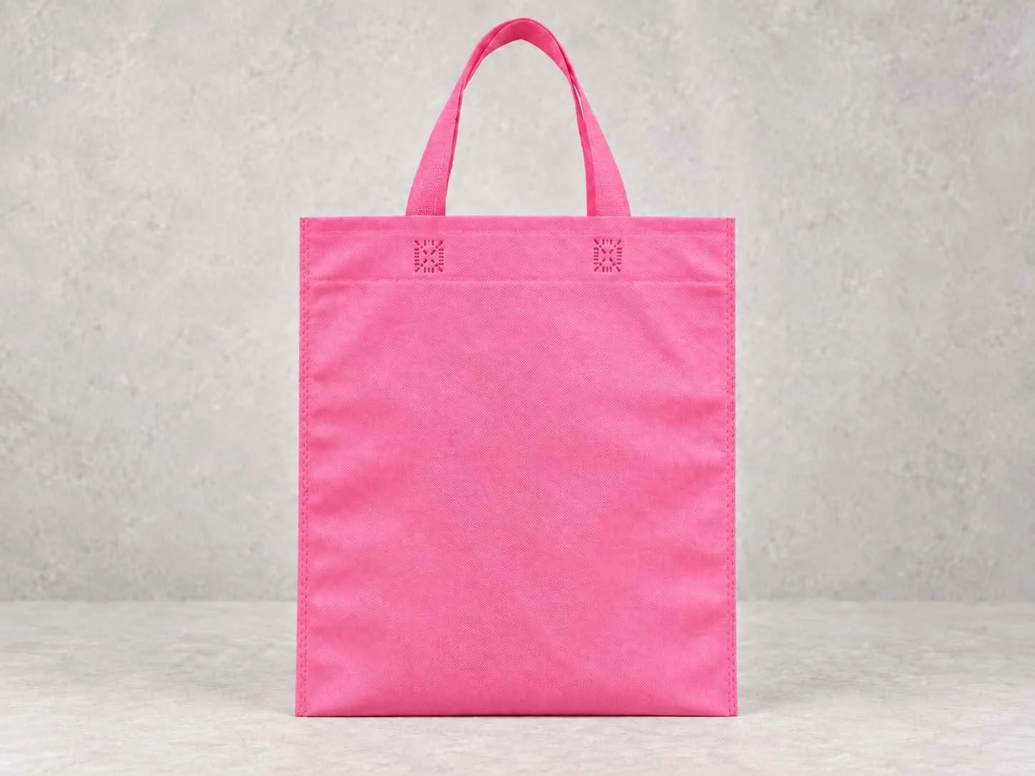 A pink reusable shopping tote bag made of fabric, standing upright on a light gray surface against a textured light gray background.