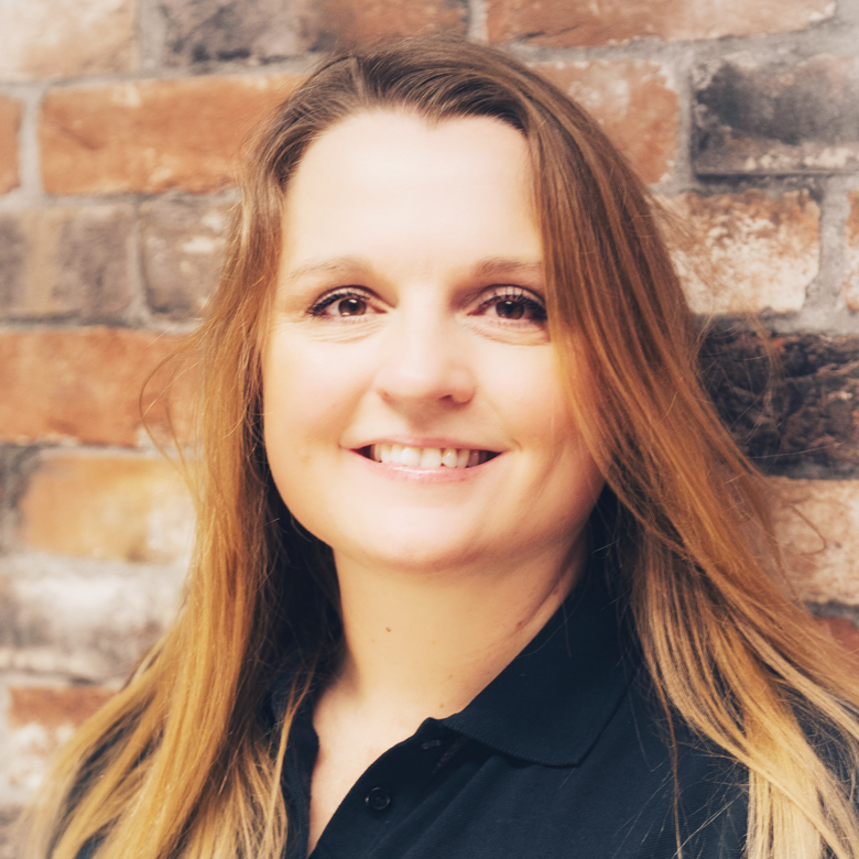 Close-up of a woman with long red hair smiling, wearing a black shirt, standing in front of a brick wall.