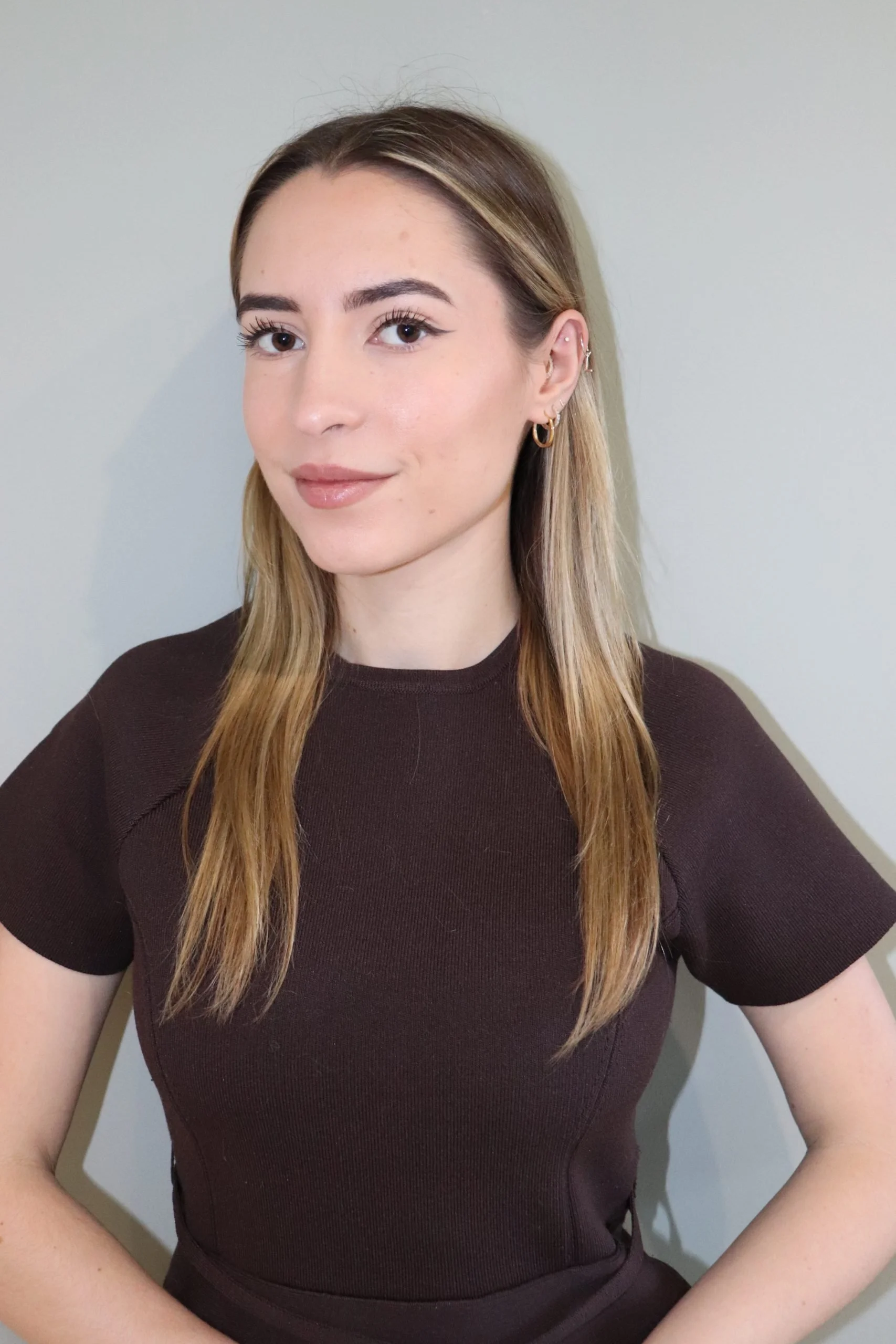 A young woman with long, wavy blonde hair wearing a dark brown t-shirt, standing against a plain, light-colored wall, looking slightly to the side and smiling softly.