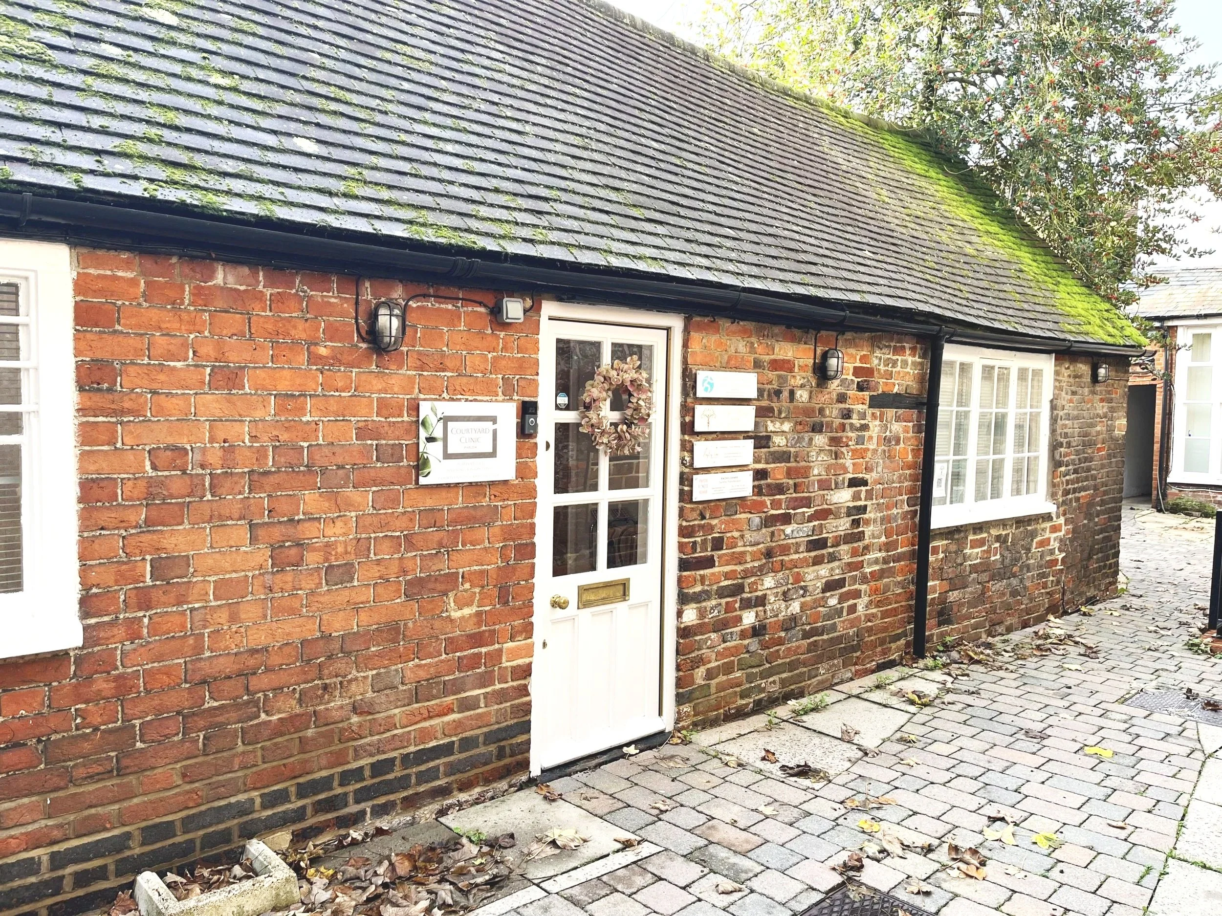 A brick building with a moss-covered sloped roof and a front door decorated with a wreath, flanked by wall-mounted outdoor lights and informational signs, with a cobblestone and concrete pathway in front.