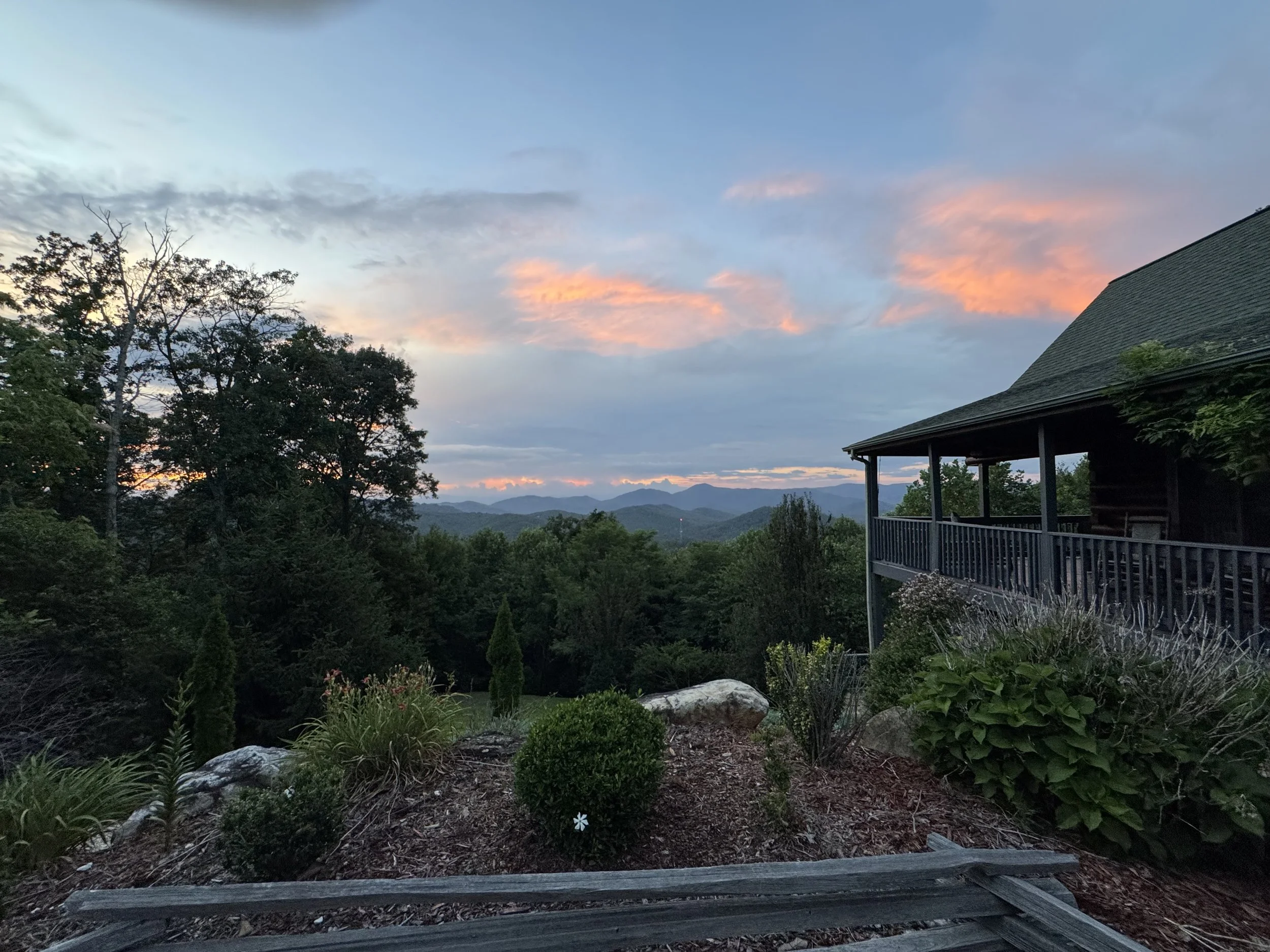 A scenic view of a sunset sky with soft pink, orange, and blue clouds over distant mountains, with a porch and lush greenery in the foreground.